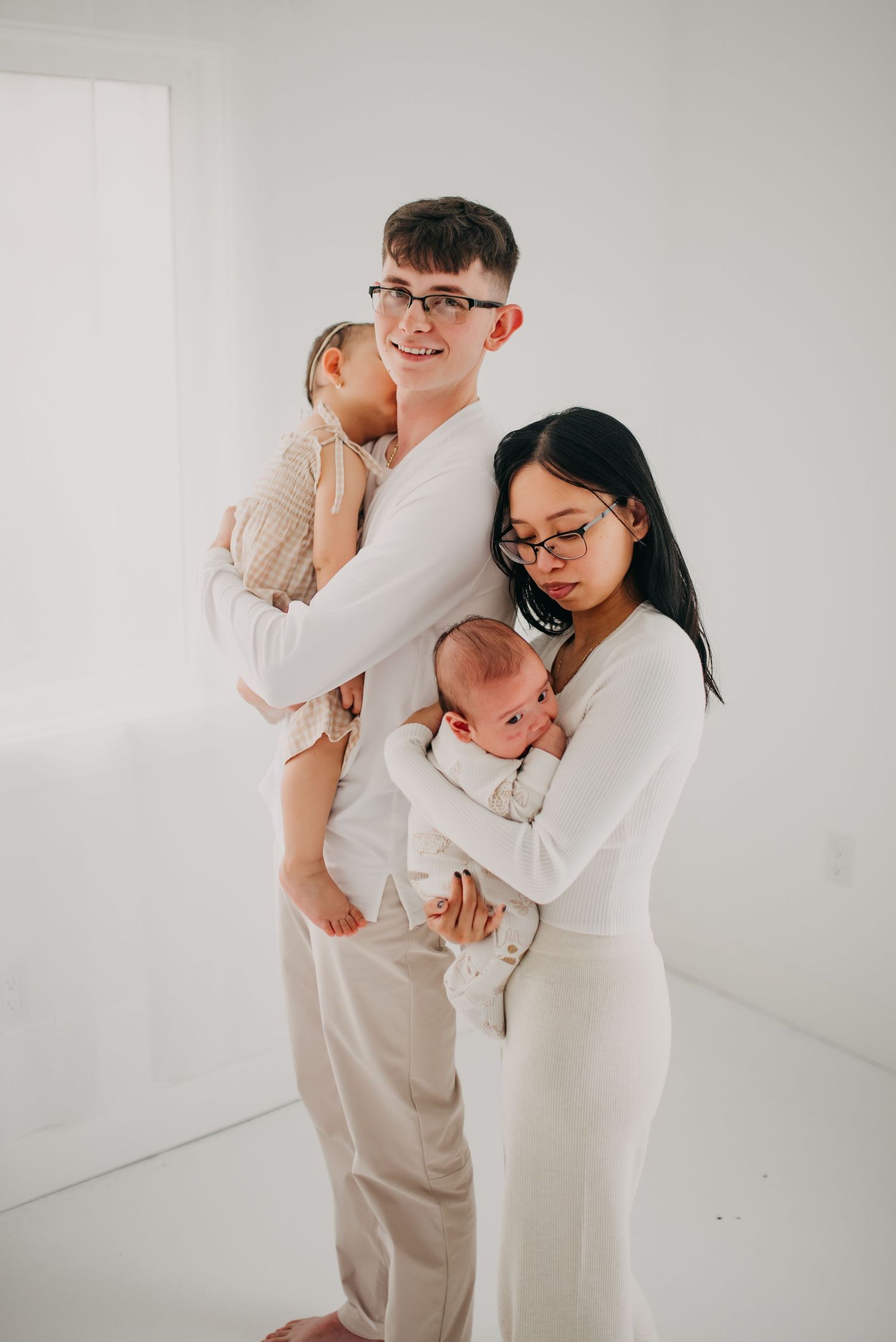Young family with toddler and newborn posing together in bright white studio.