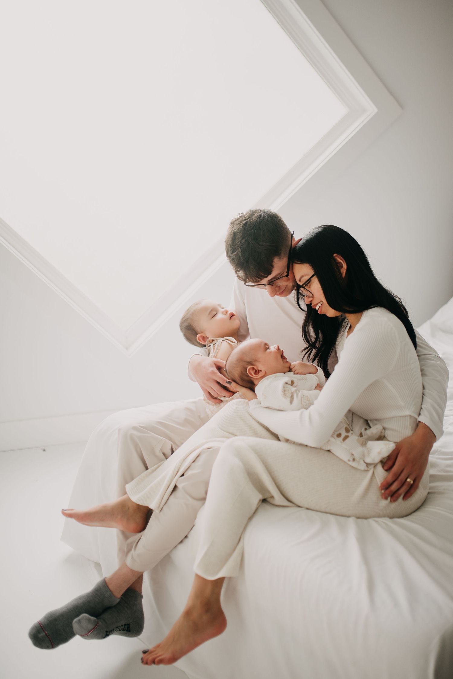 Parents sitting on white bed gazing lovingly at twin newborn babies in a bright, airy room.