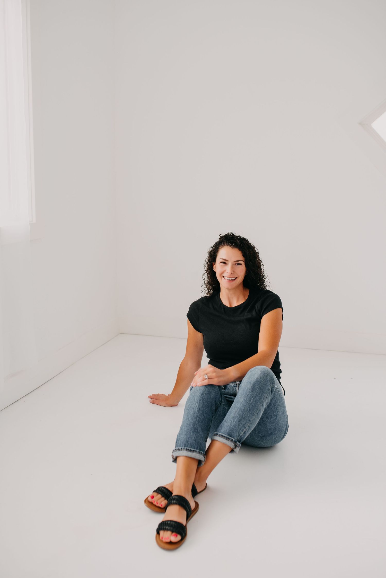 Smiling woman with curly hair sitting relaxed on floor in minimalist white studio.