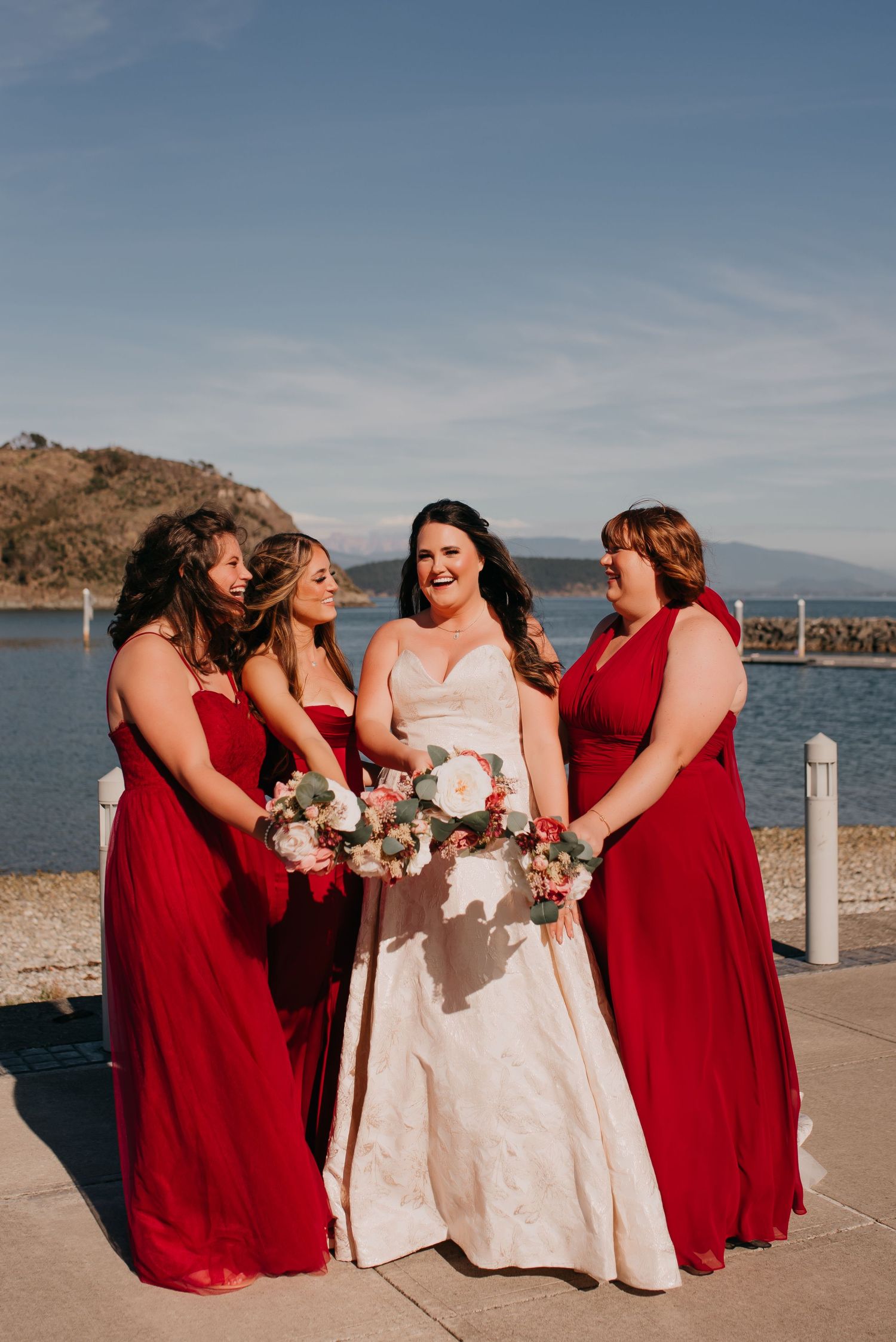 Bride in white gown poses with three bridesmaids in red dresses by a waterfront on a sunny wedding day.