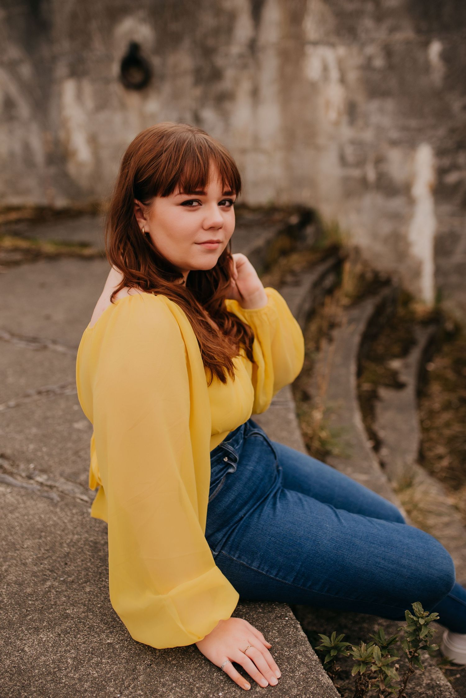 Young woman with auburn hair in yellow blouse and jeans sits on stone wall for senior portrait.