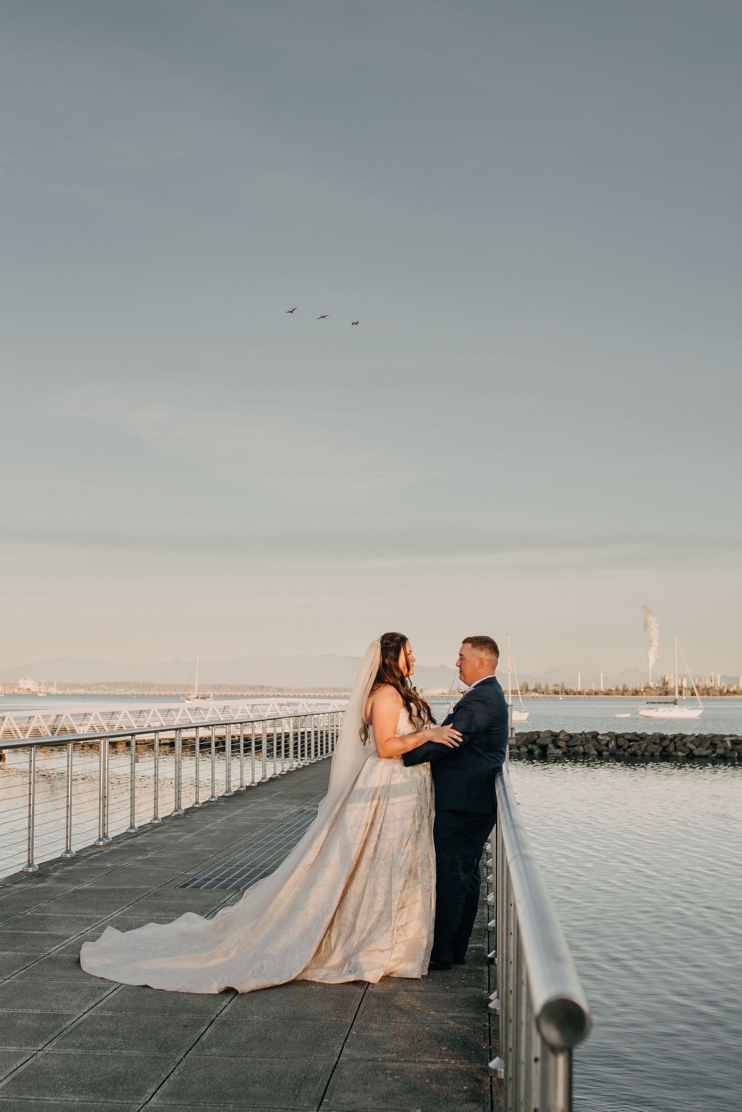 Bride and groom holding hands on a waterfront pier at golden hour with calm water behind.