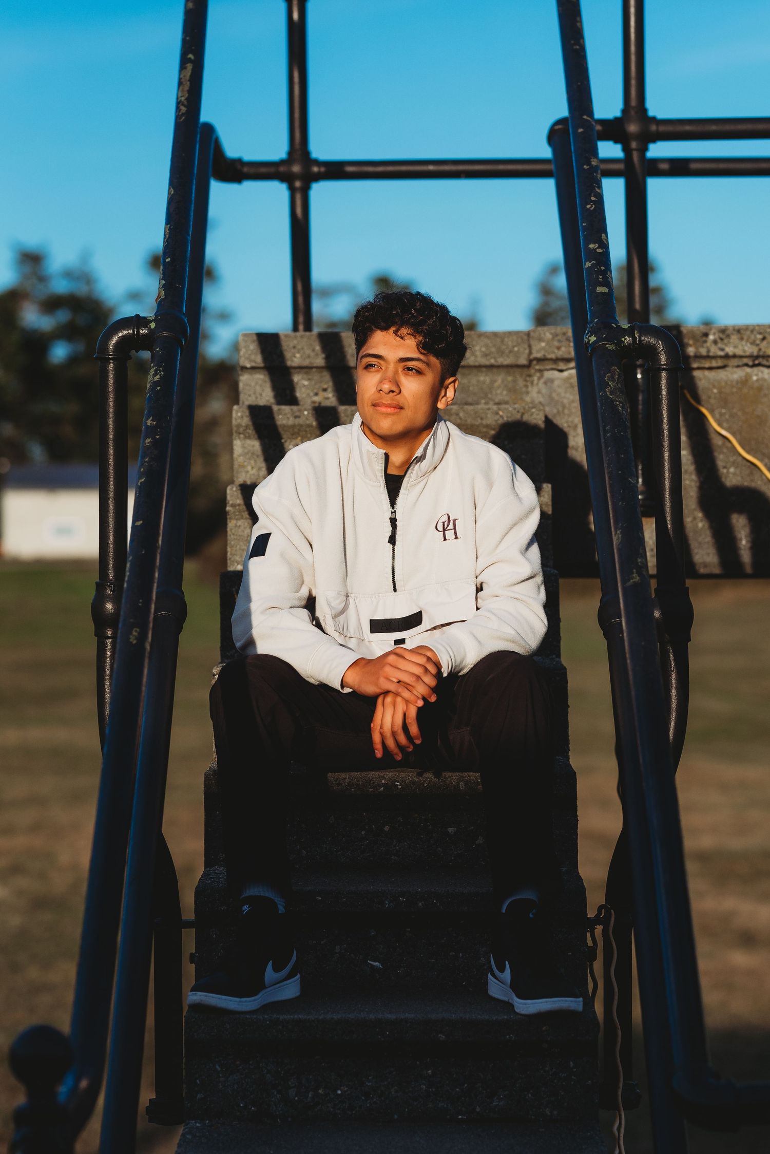 Young man in white jacket sitting on bleacher steps at an outdoor sports field during golden hour.