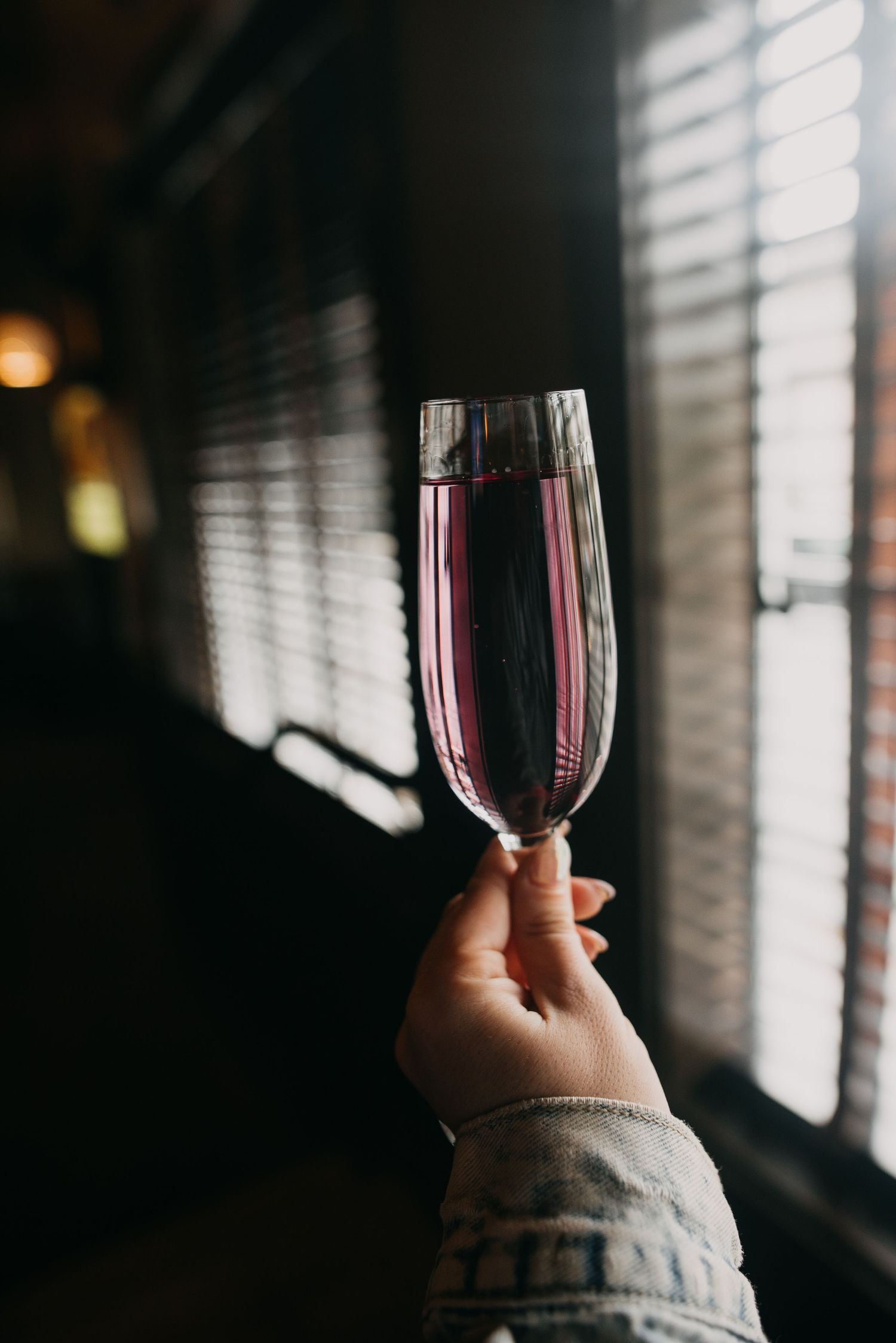 Hand holding a champagne flute with pink sparkling wine near window with venetian blinds.