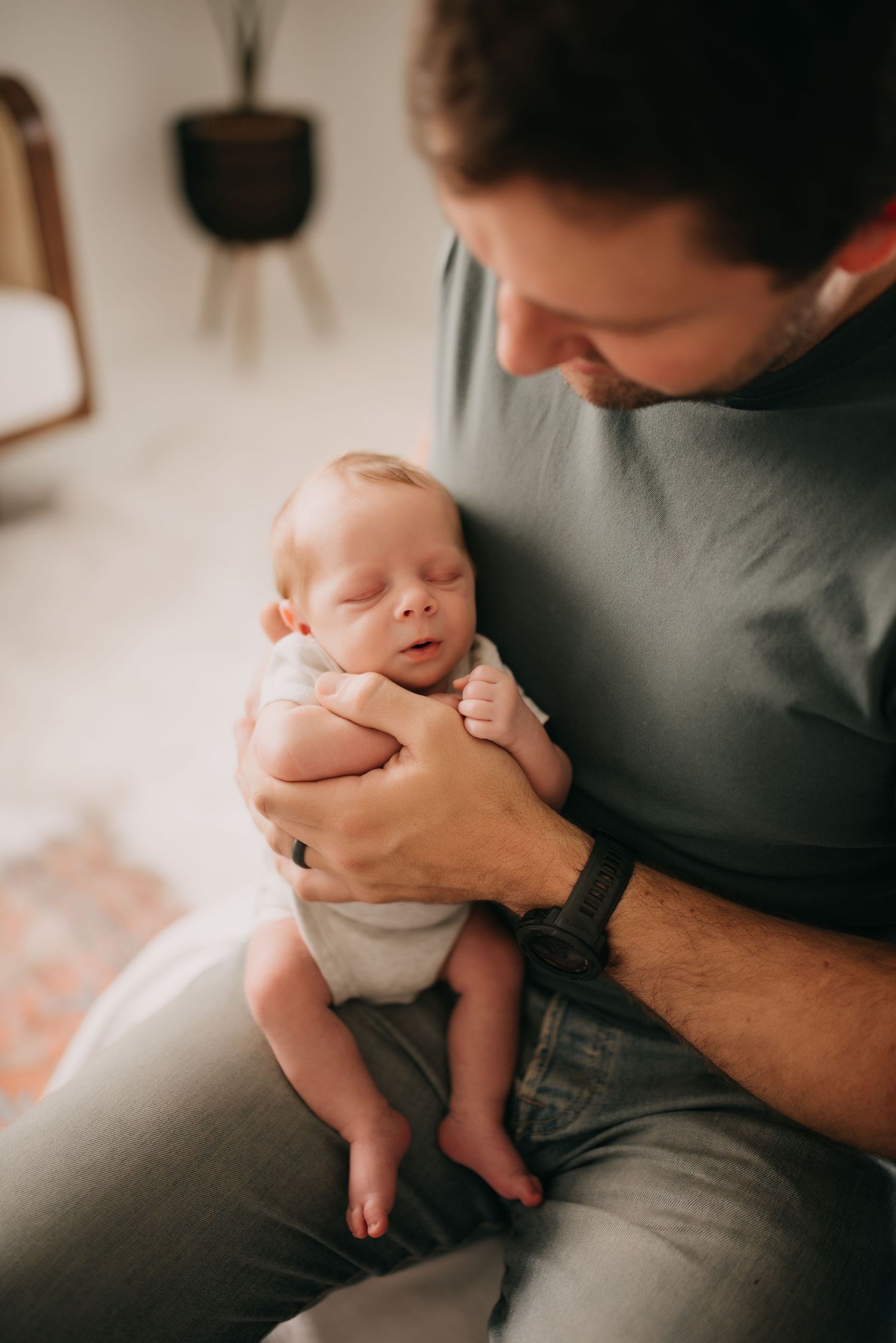 Father in teal shirt tenderly holds sleeping newborn baby against his chest indoors.