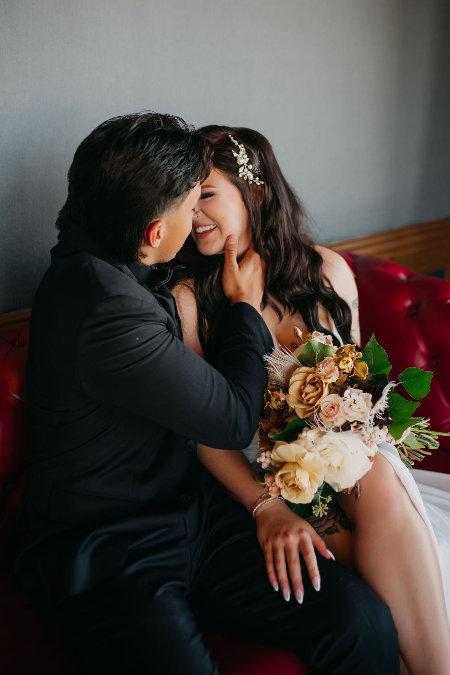 Laughing bride in wedding dress holds bouquet while sitting with groom on red velvet sofa.