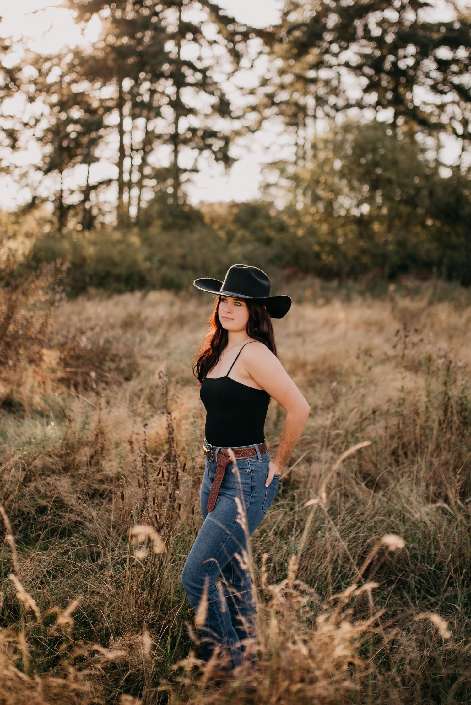 Young woman in black top and wide-brim hat poses in sun-lit golden field at sunset.