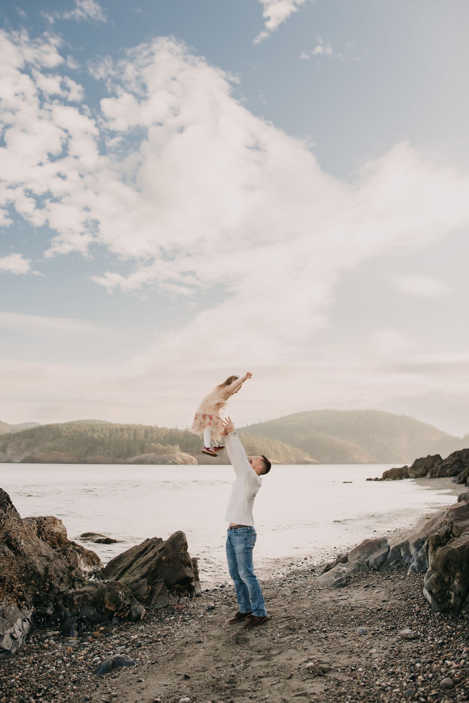 Father tossing young child in the air on a rocky beach with mountains and cloudy sky in background.