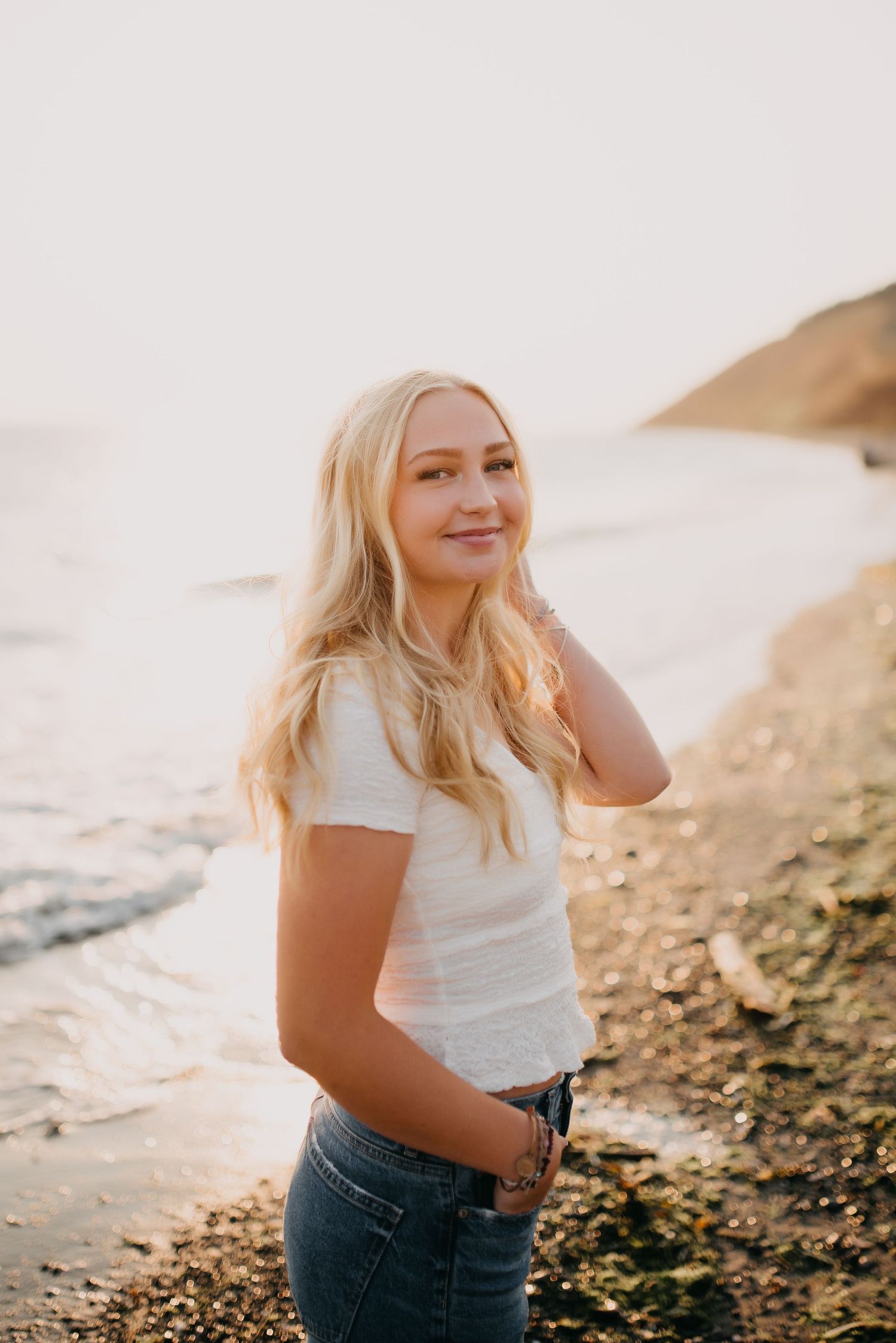 Smiling blonde woman in white top and jeans poses on a sunlit rocky beach with ocean waves behind her.
