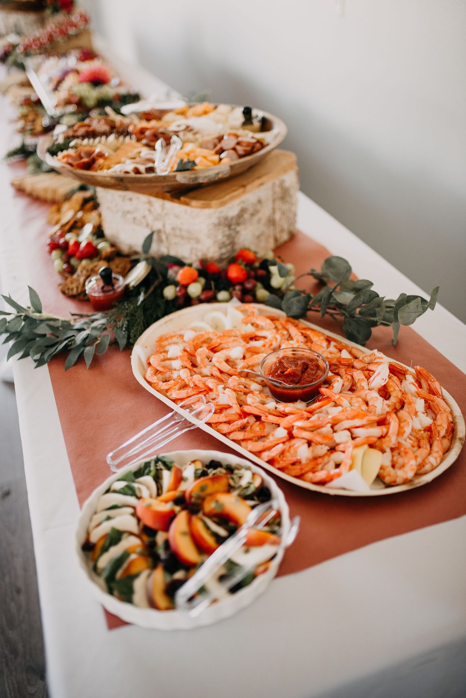 Wedding reception food table featuring shrimp cocktail, charcuterie board, and fresh salad with floral garland decor.