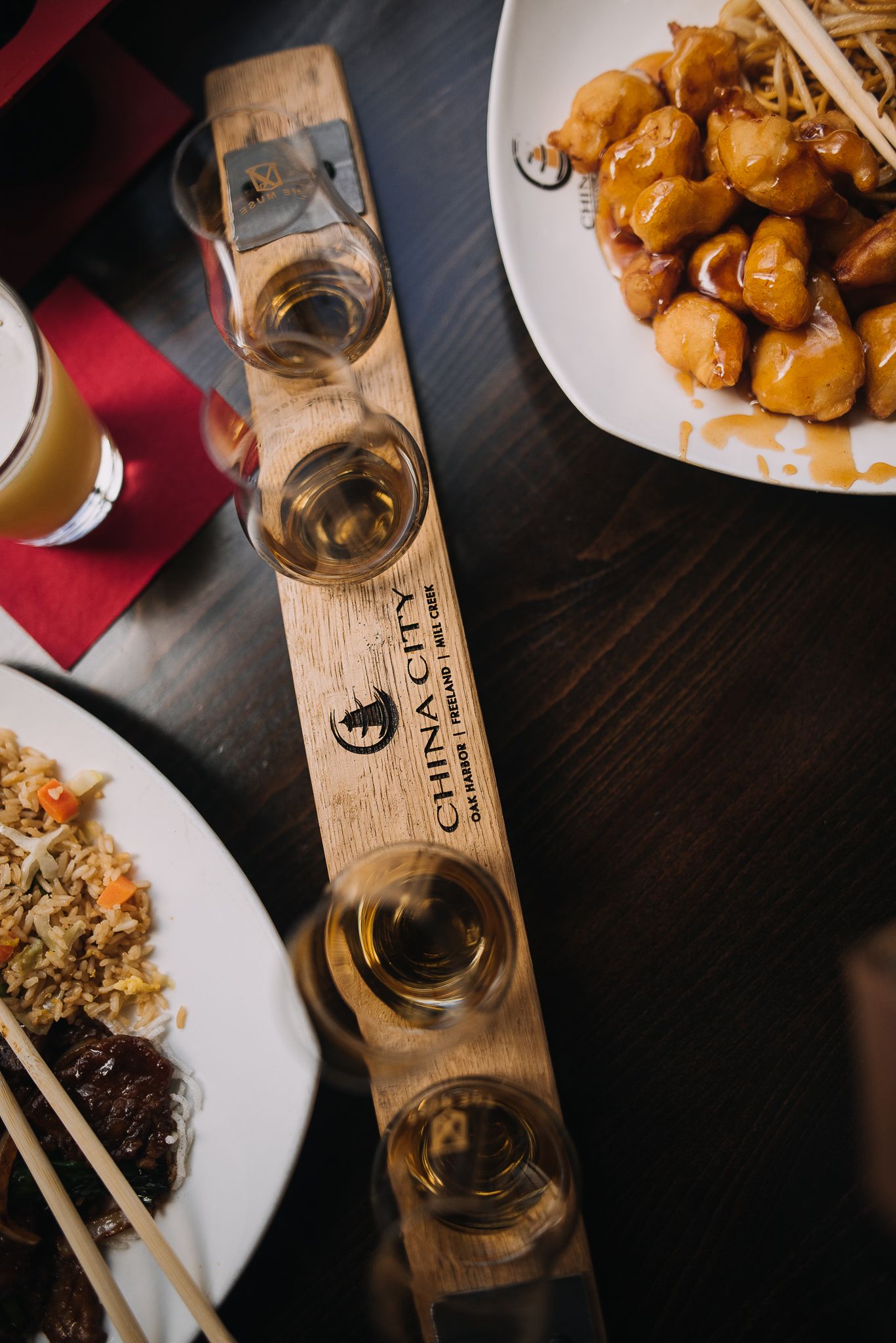 China City wooden whiskey flight paddle with shot glasses beside Asian dishes on a dark table.