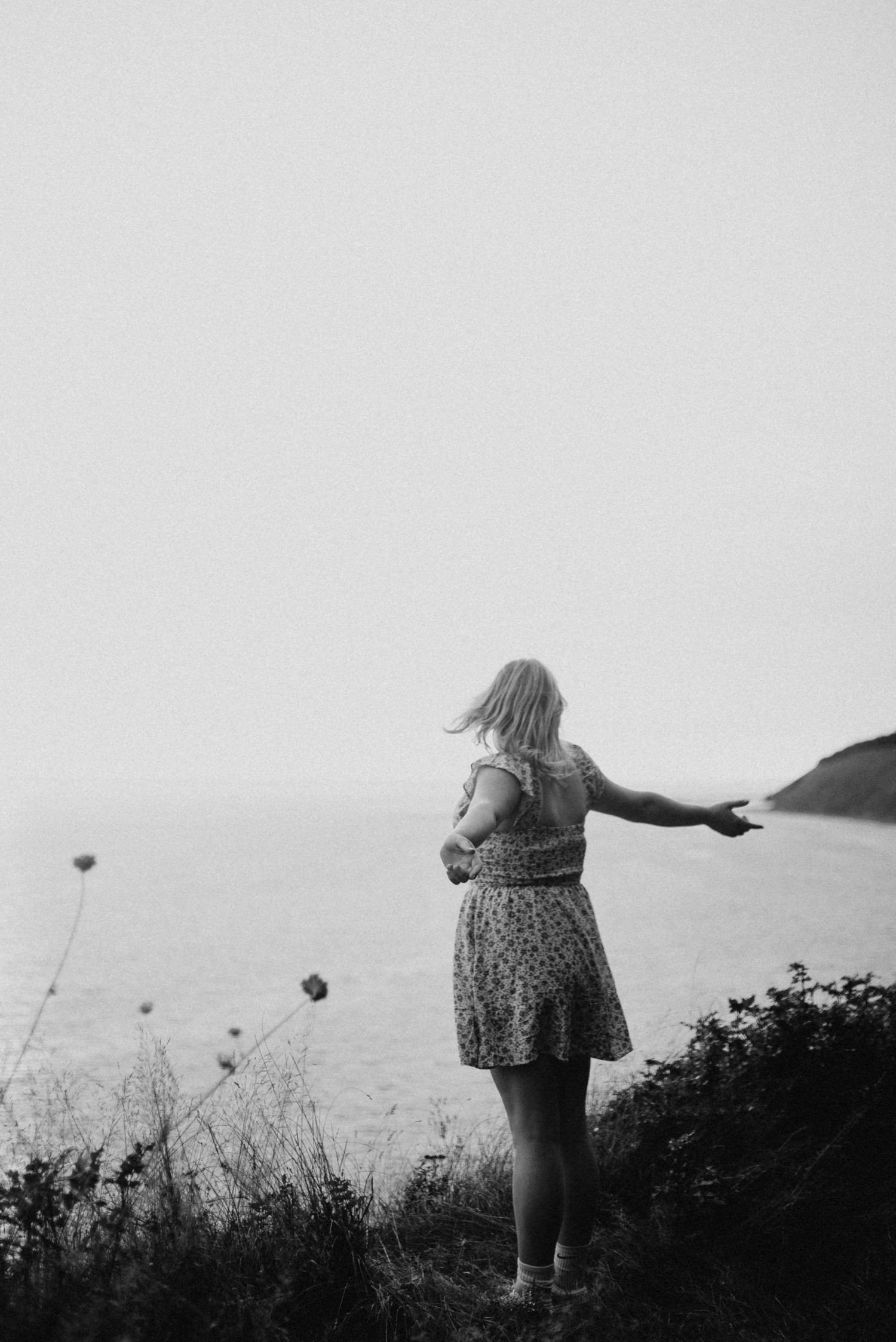 Woman in floral dress stands on coastal cliff with arms outstretched overlooking the ocean.