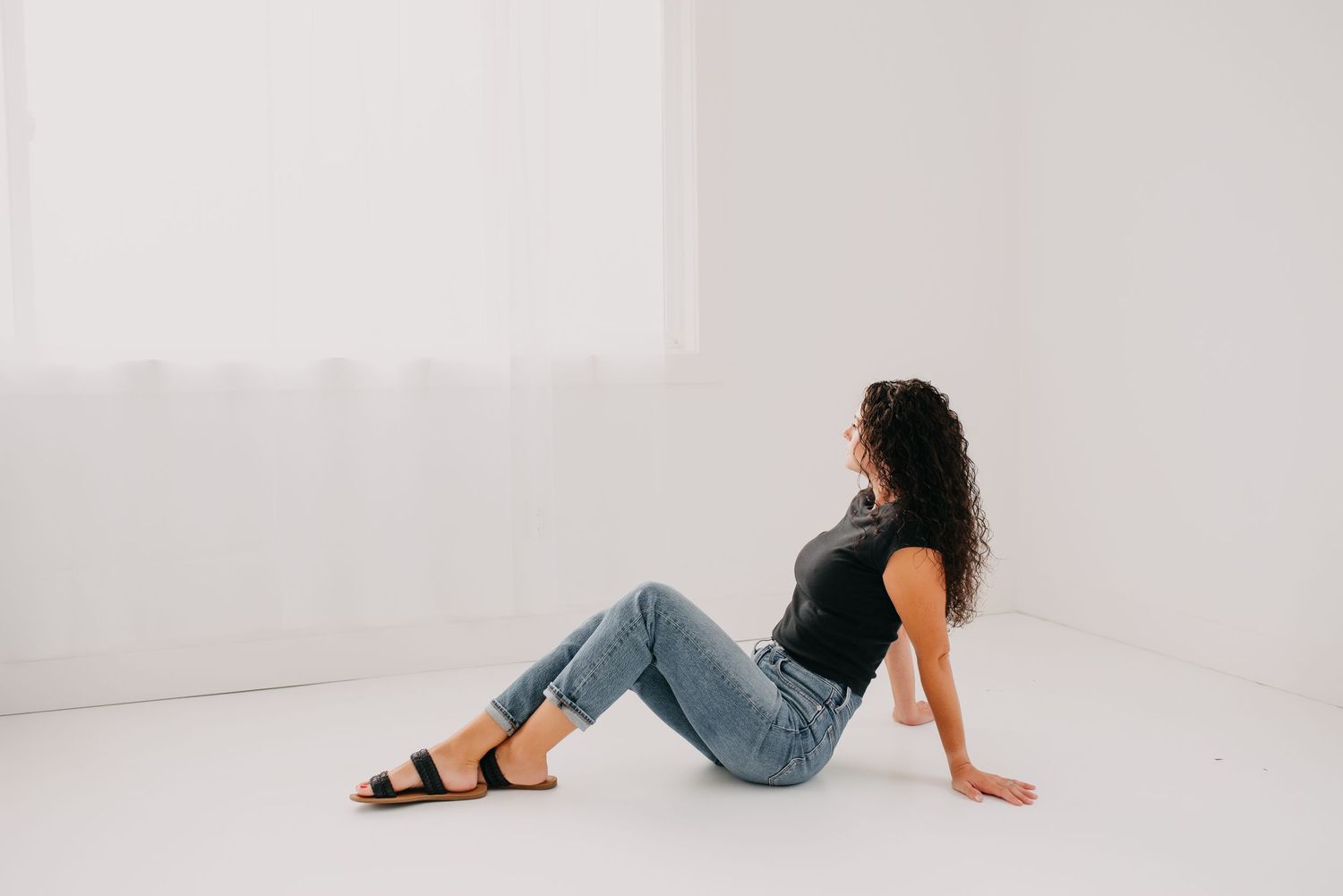 Young woman with curly hair sitting casually on floor in black top and jeans against white background.