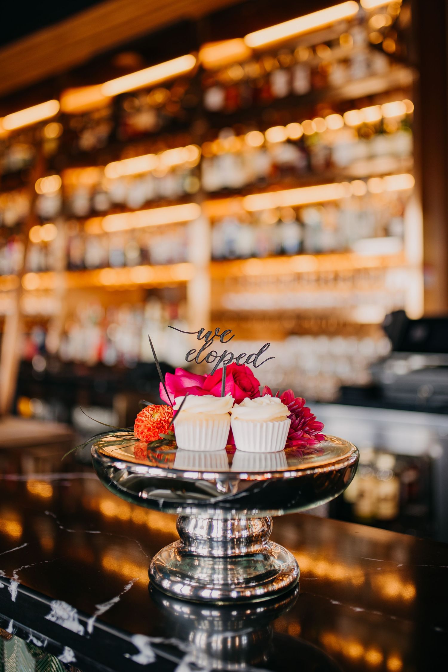 Wedding cupcakes with 'we eloped' topper on silver stand at a bar with warm bokeh lights.