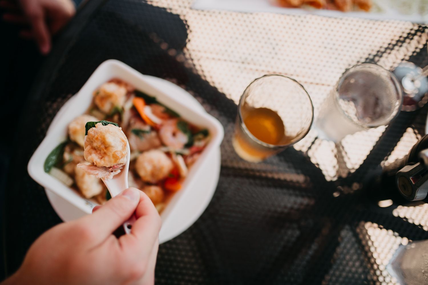 Hand holding a spoon with a shrimp over a bowl of stir-fry vegetables at an outdoor restaurant table.