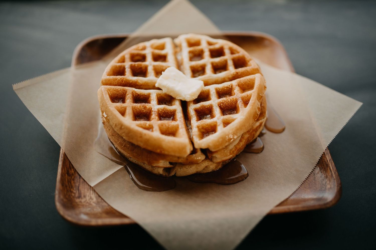 Golden waffle stack with butter pat and maple syrup served on parchment paper on wooden plate.
