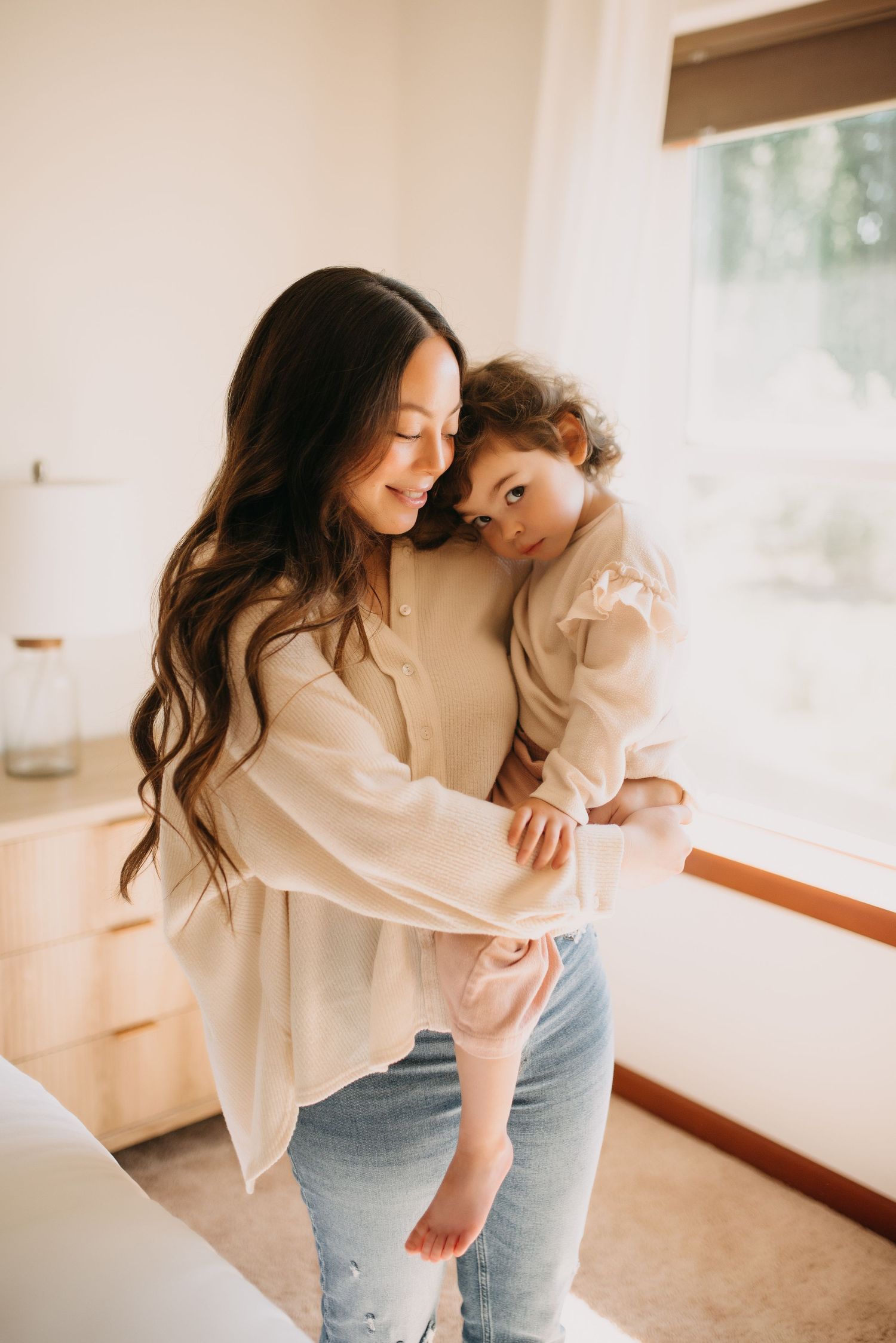 Dark-haired mother in cream blouse tenderly holding toddler near bright window in warm home.