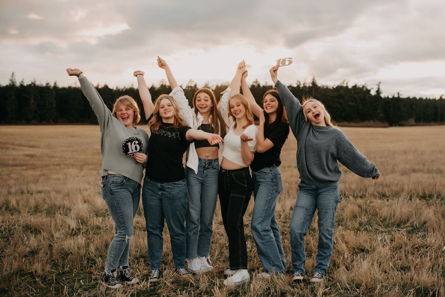 Group of six teenage girls celebrate with raised arms in a golden field at dusk.
