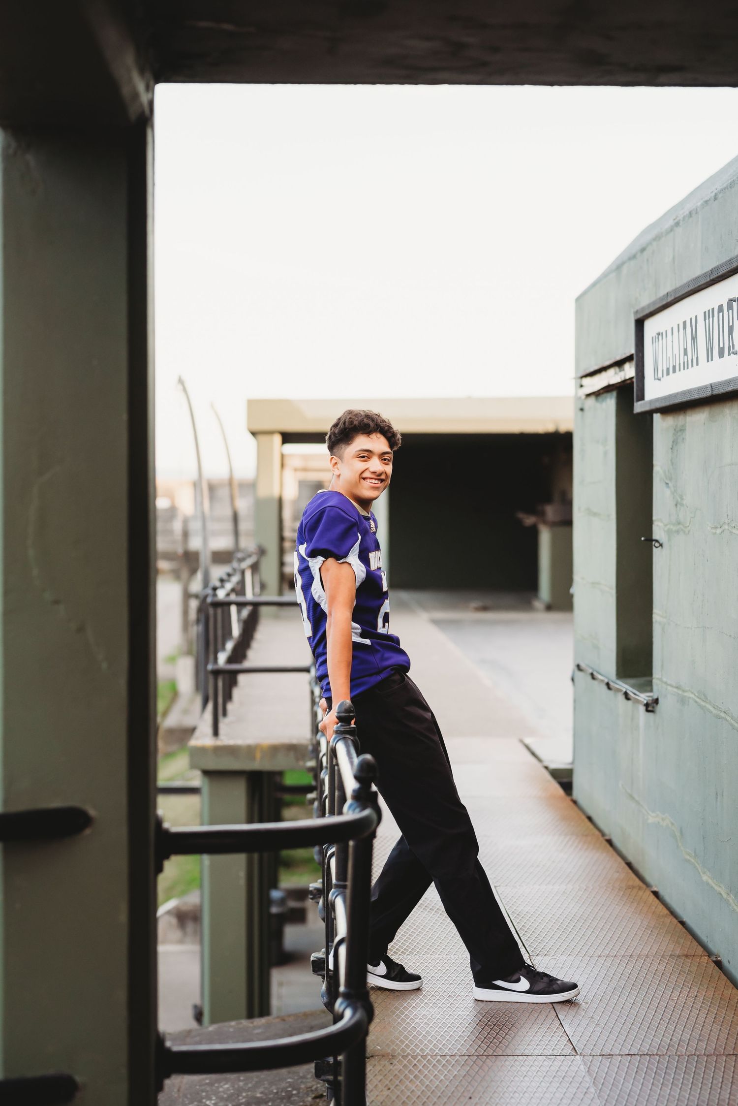 Smiling young man in purple football jersey leaning on railing at a stadium entrance.