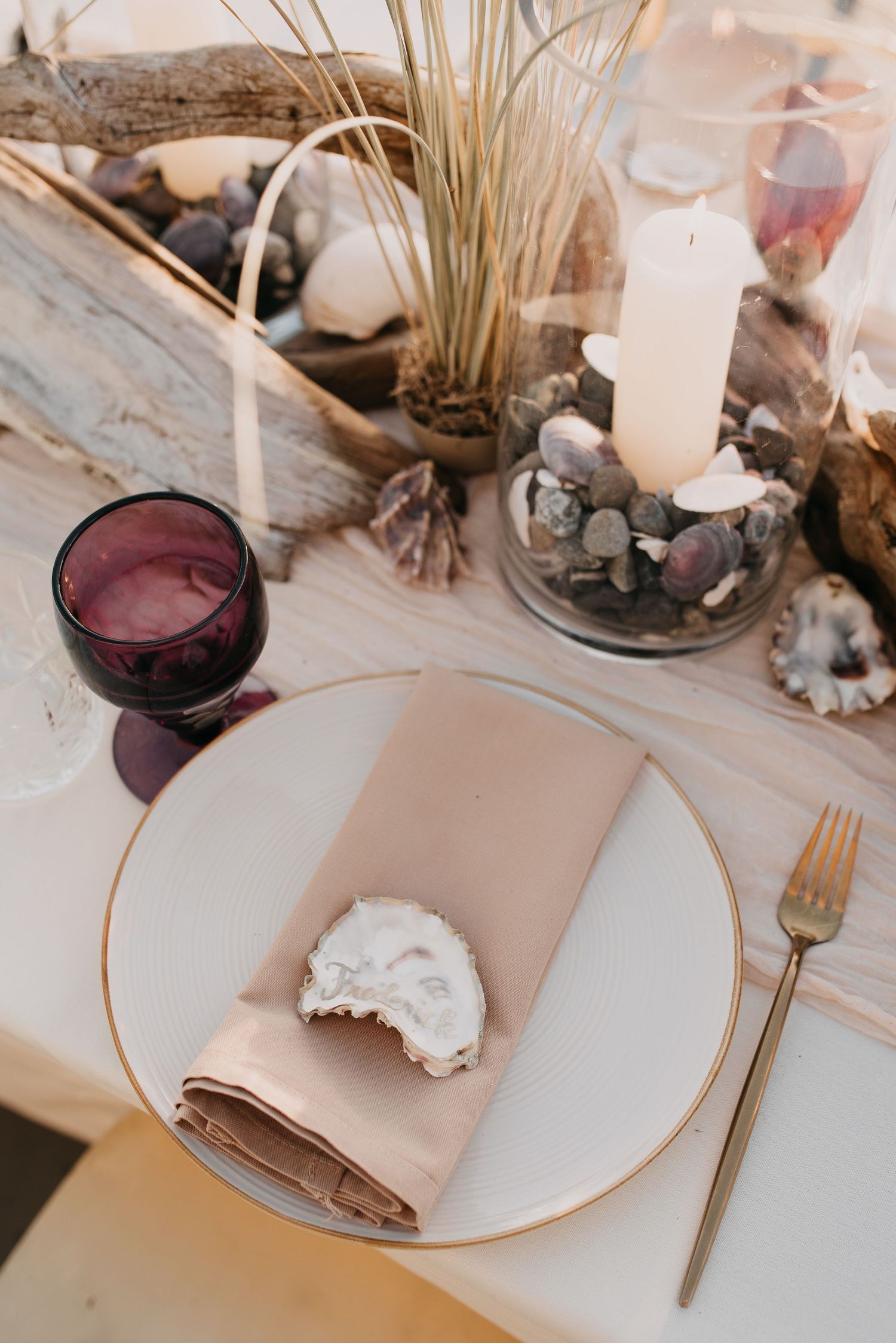 Close-up of a wedding place setting with a taupe napkin, oyster shell accent, and purple goblet.