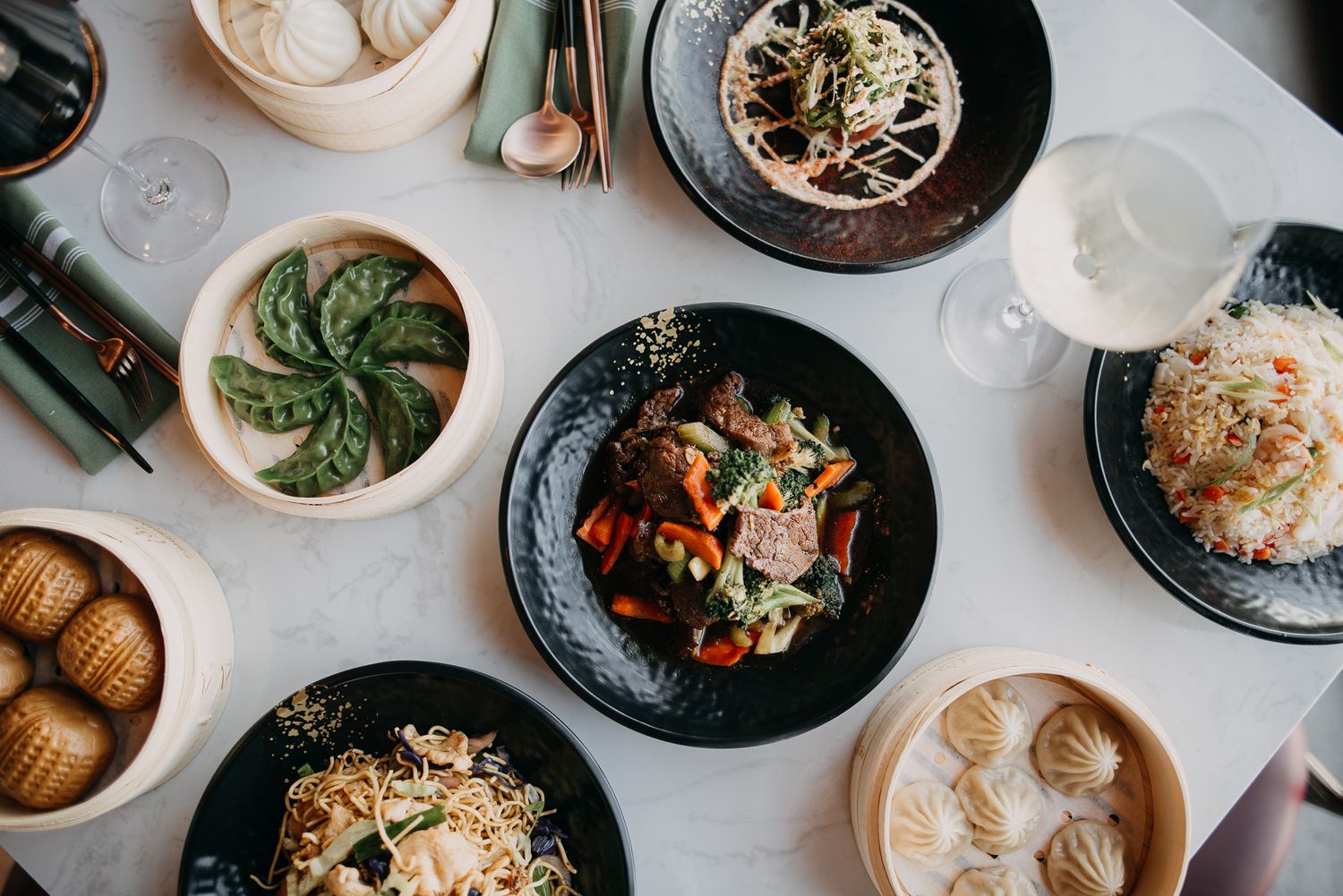 Overhead view of Asian dim sum spread with dumplings, noodles, and stir fry on dark plates.