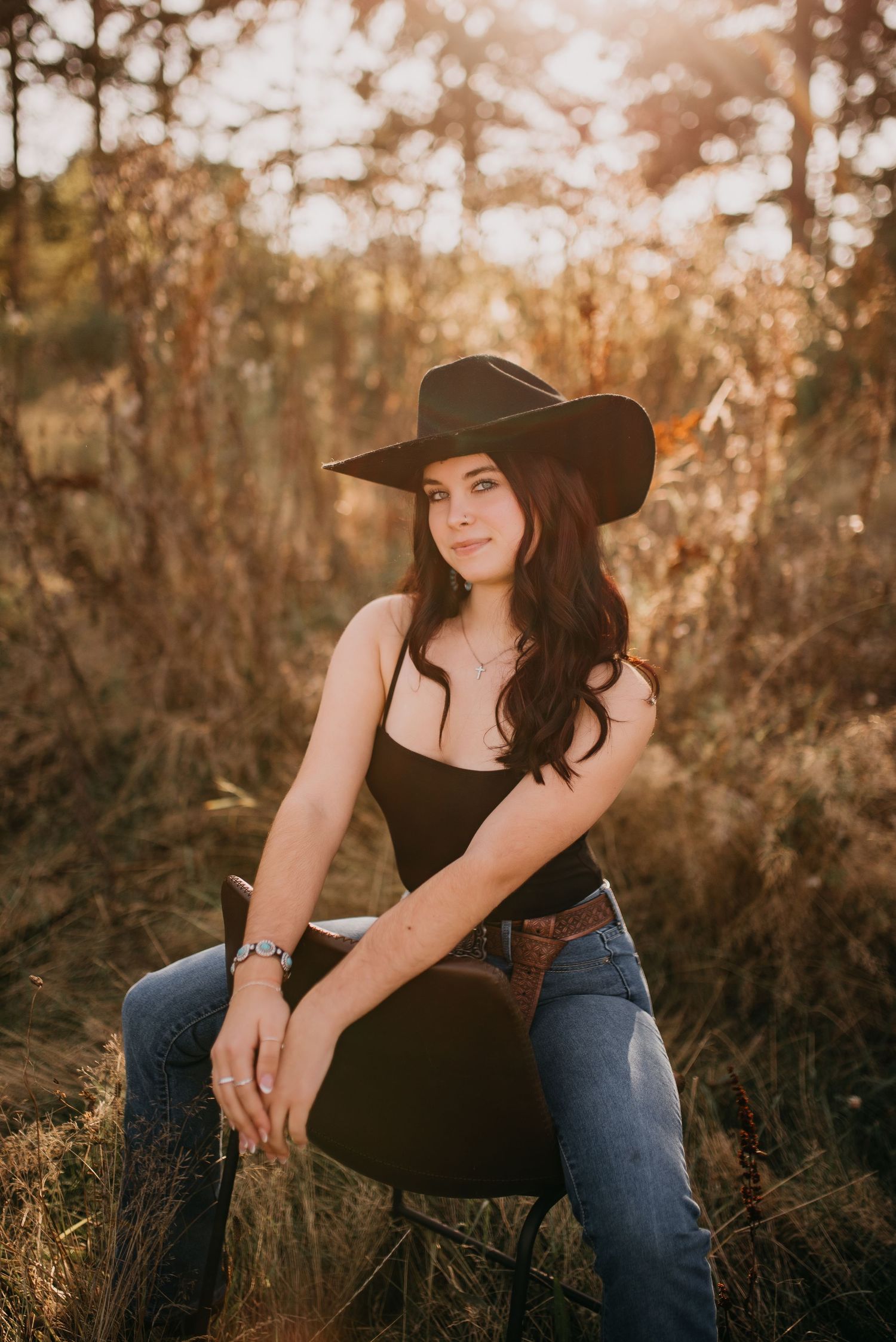 Young woman in black hat and bodysuit sits on chair in golden sunlit field for senior portrait.