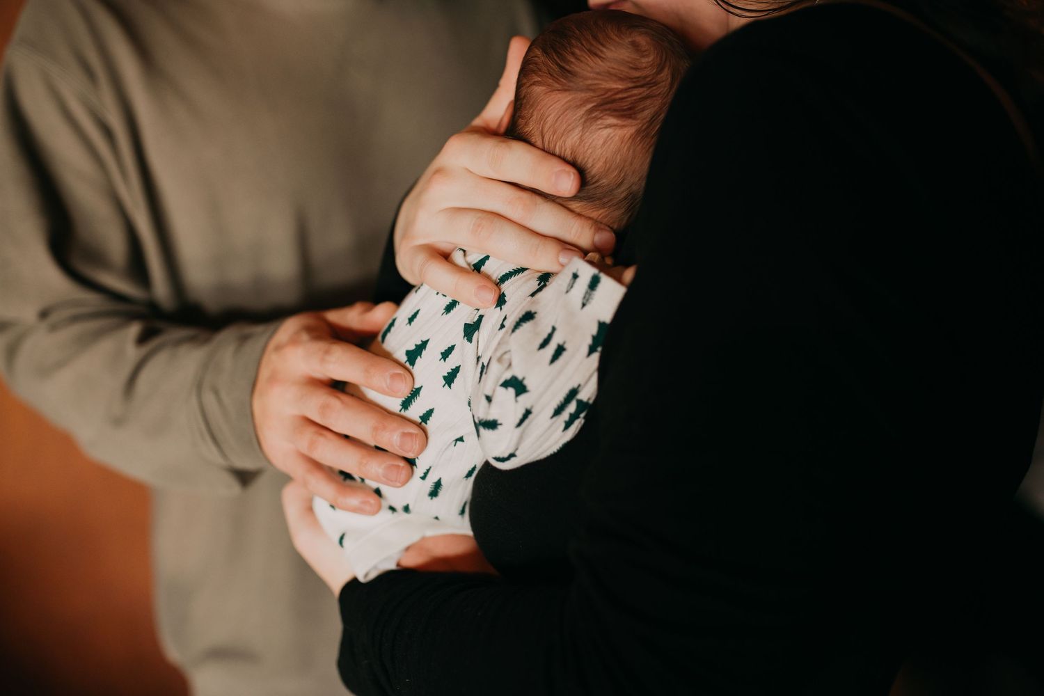 Parents cradling newborn baby dressed in white patterned onesie in a warm, intimate close-up photo.