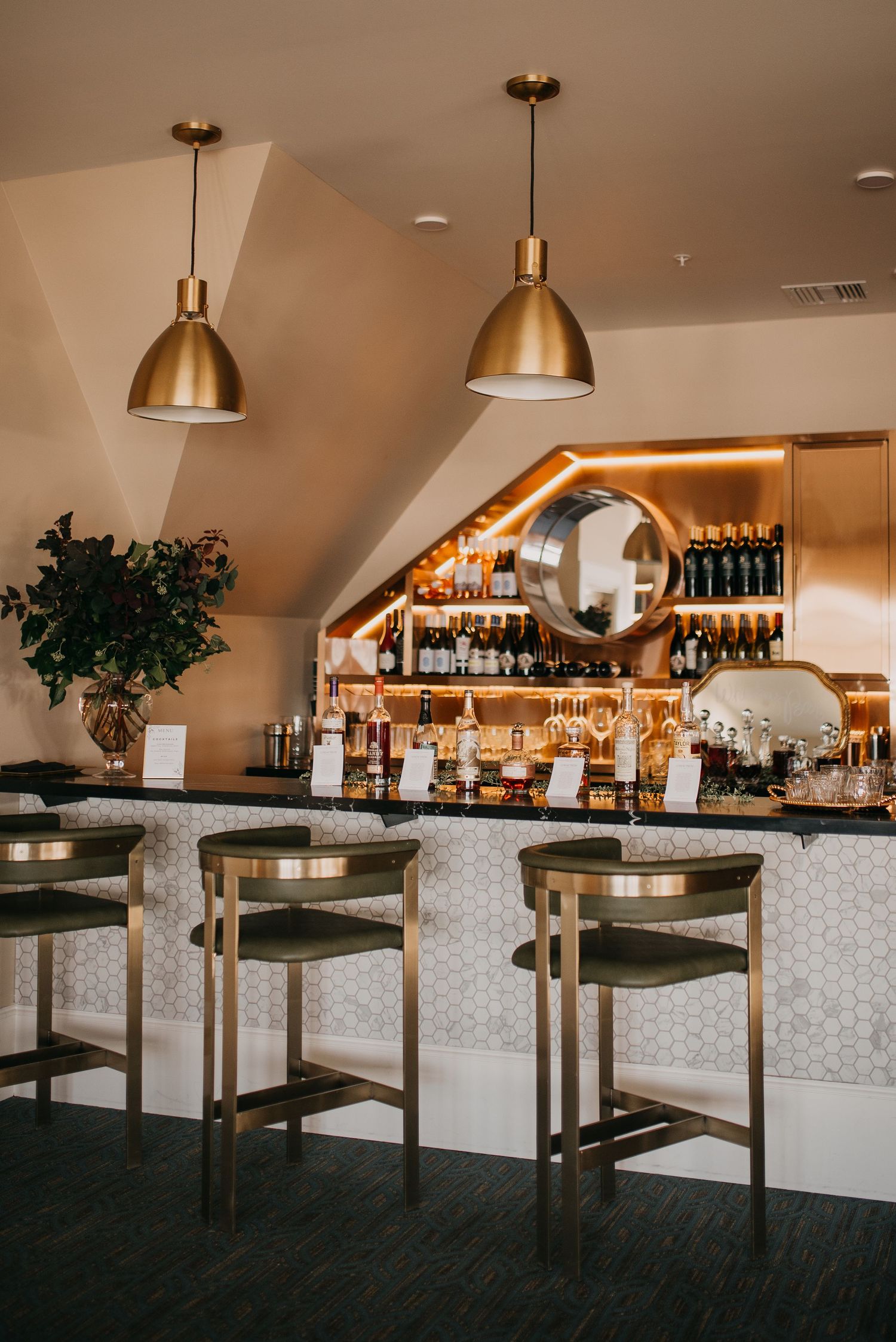 Elegant wedding venue bar with gold pendant lights, green velvet stools, and backlit liquor display.