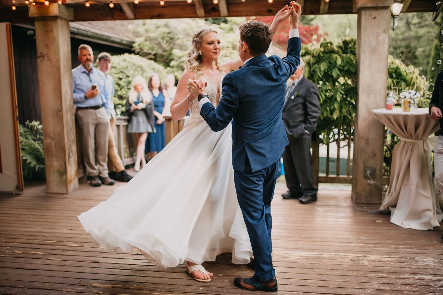 Bride in flowing white gown dances with groom in blue suit on a wooden deck at outdoor reception.