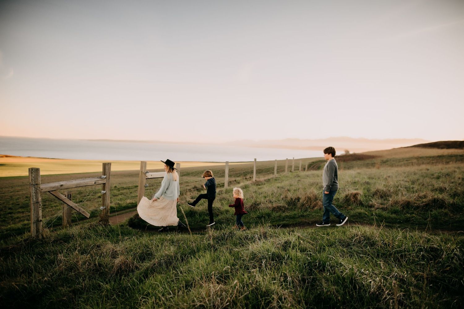 Family walking together through green countryside field at golden hour near wooden fence.