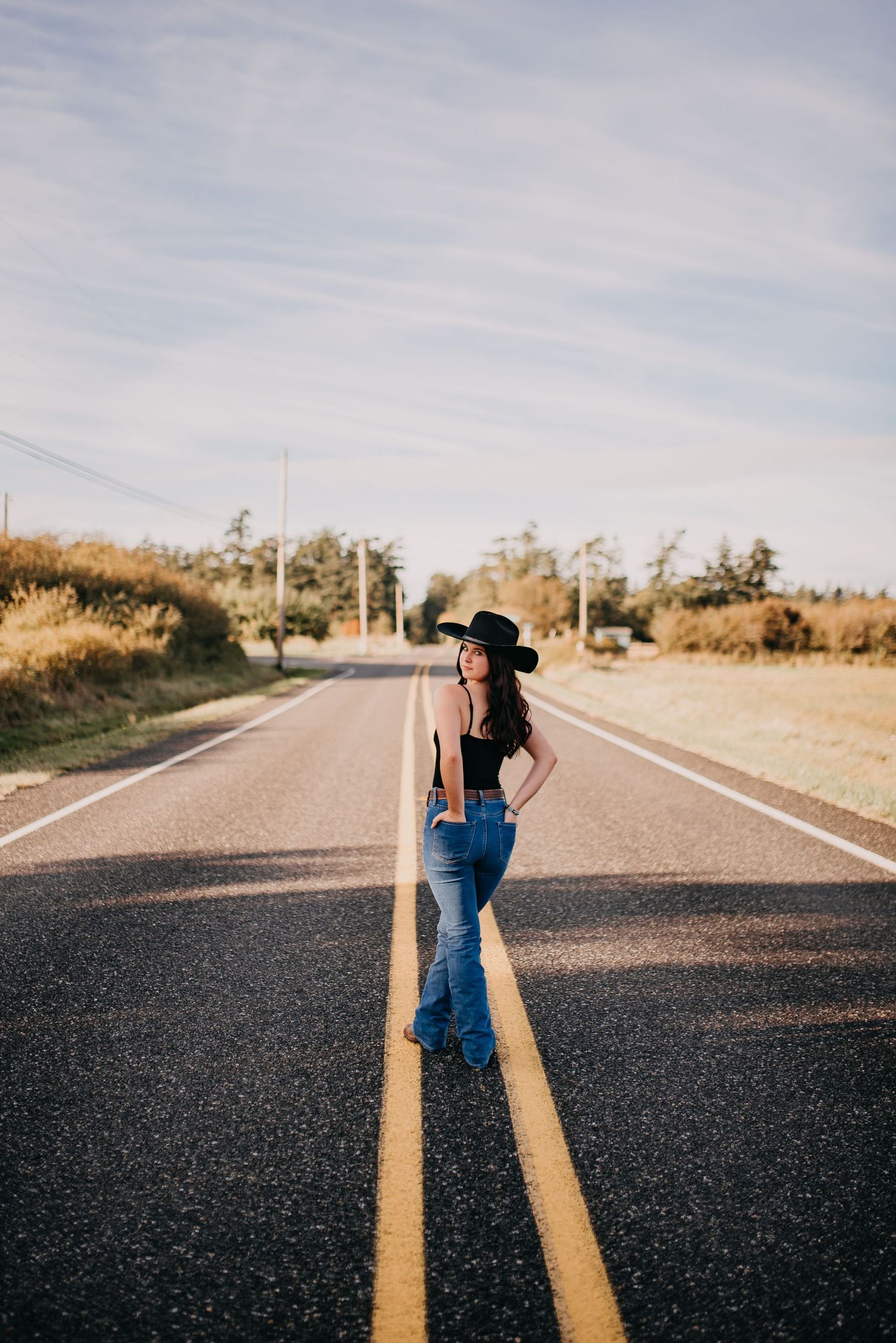 Young woman in black hat and flare jeans stands confidently on empty rural road at golden hour.