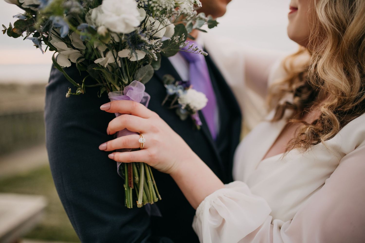 Close-up of bride and groom holding white and blue floral bouquet with purple ribbon at wedding ceremony.