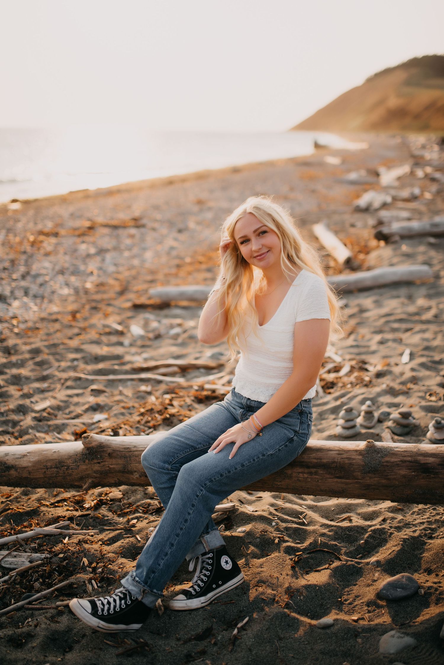 Blonde woman in white tee, jeans, and Converse sits on driftwood at a sandy beach during golden hour.