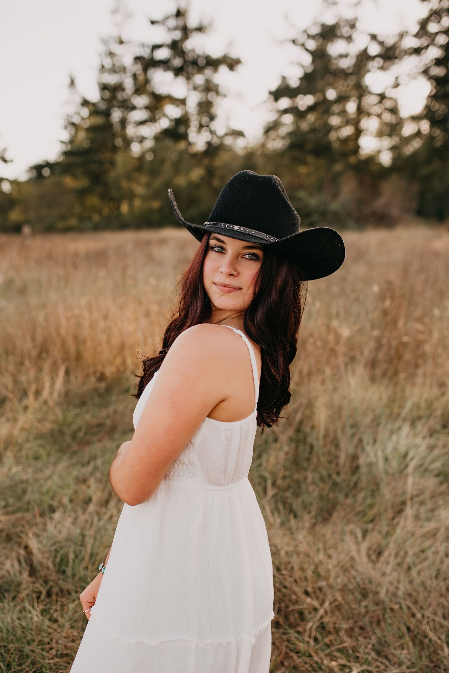 Young woman in white dress and black cowboy hat poses in golden field during sunset.