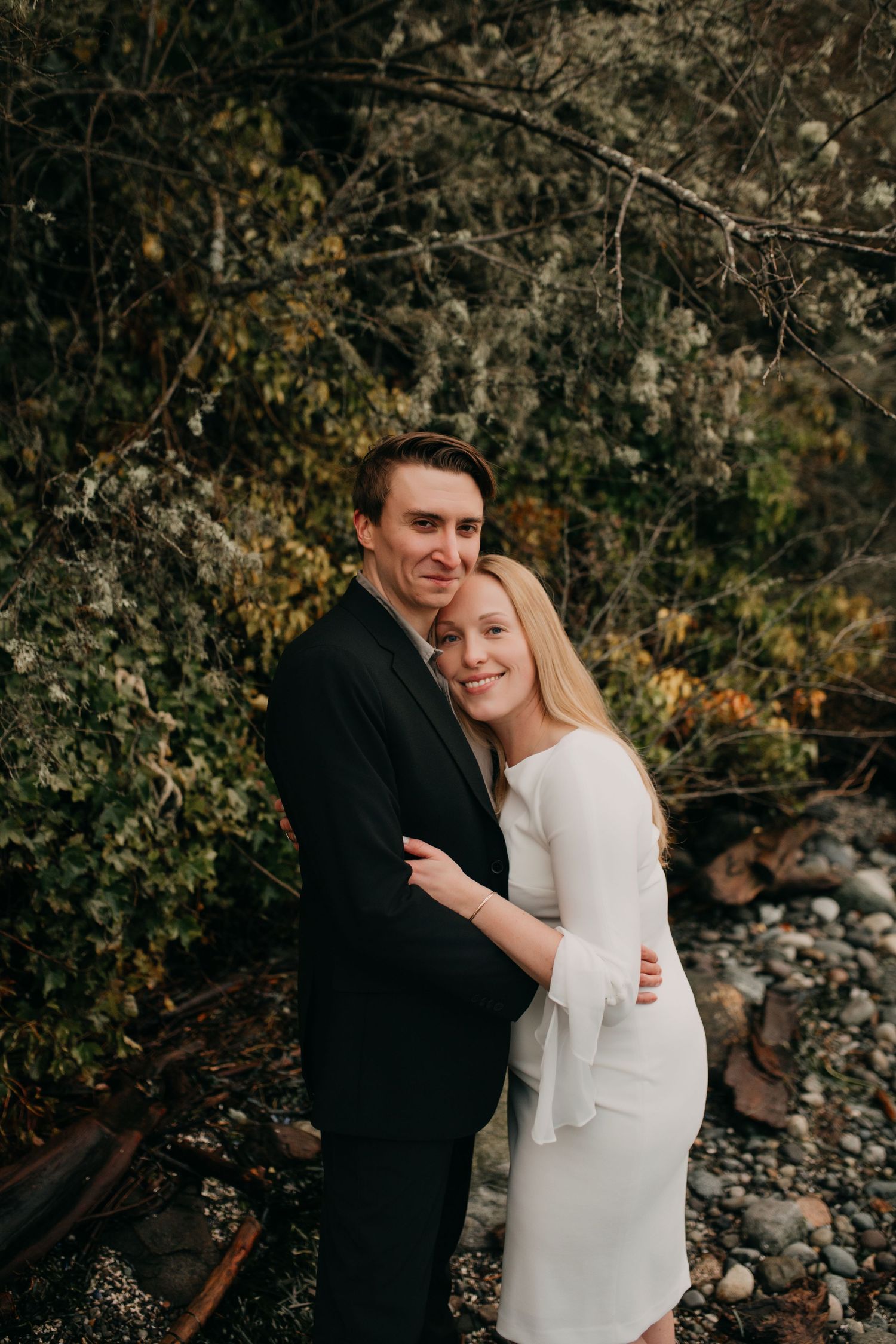 Couple embraces tenderly in front of lush greenery and rocky shoreline during an outdoor engagement session.
