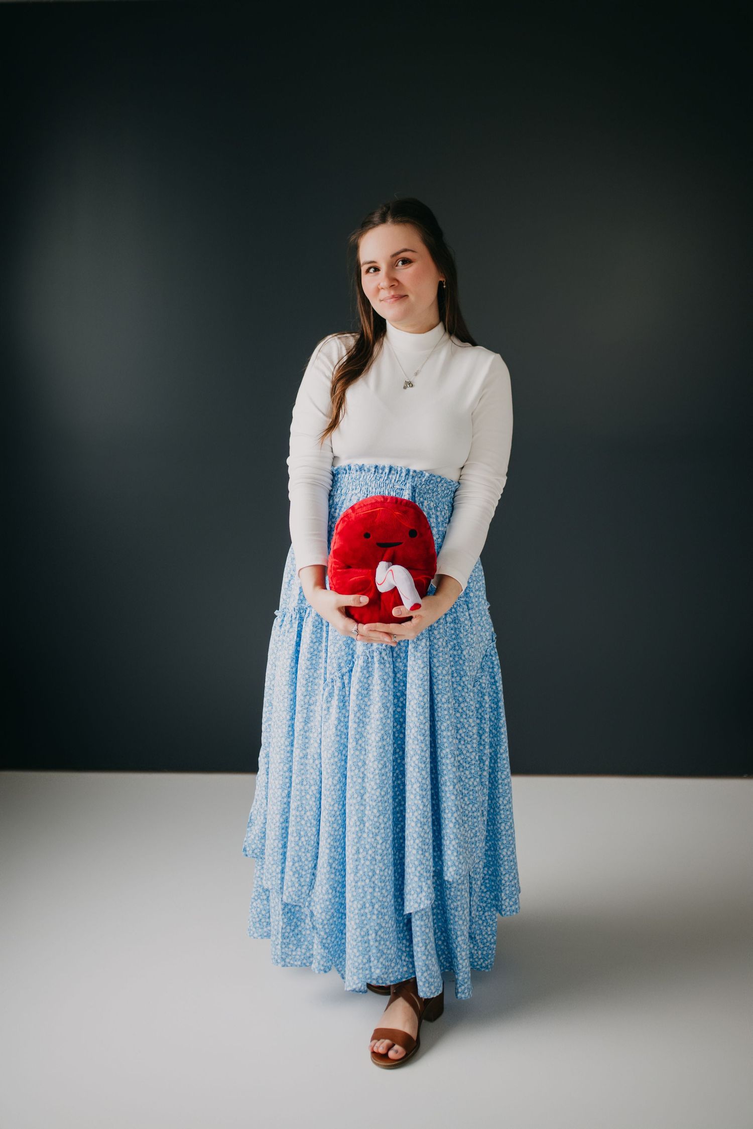 Pregnant woman in white crop top and blue floral maxi skirt holding red stuffed animal against dark background.
