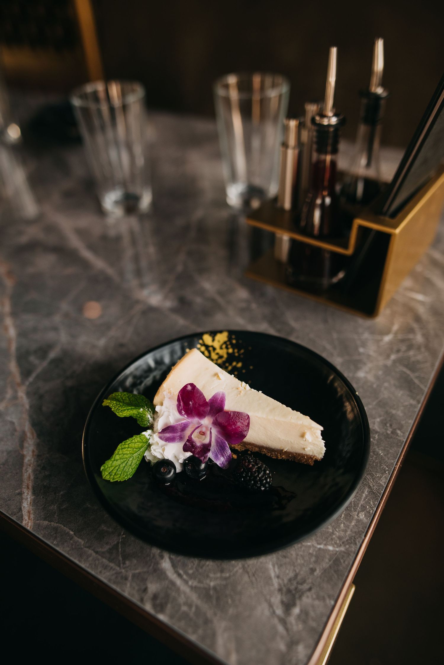 Elegant cheesecake slice garnished with purple orchid, blackberries, and mint on a black plate.