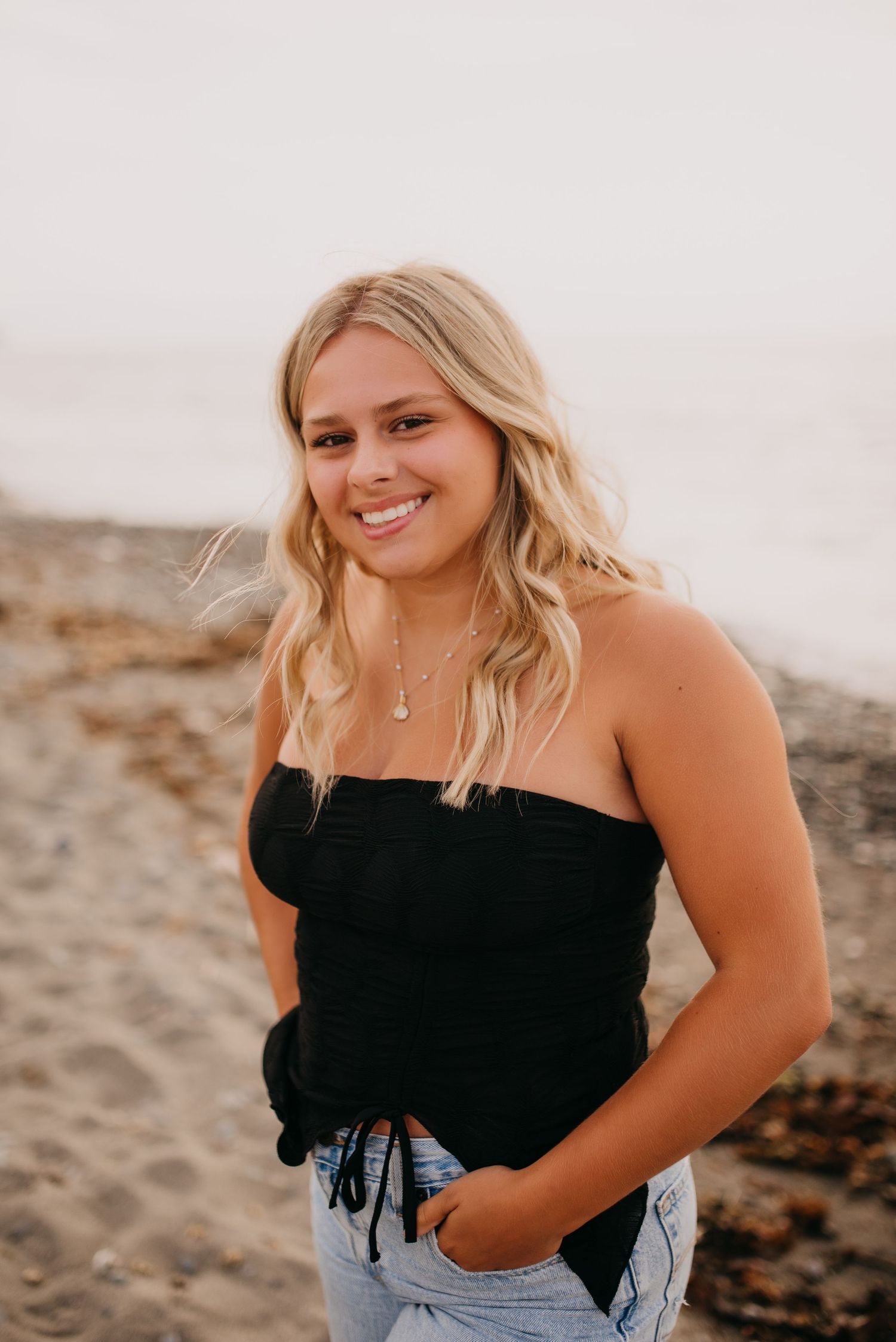 Blonde teen girl in black strapless top smiling on sandy beach during senior portrait session.