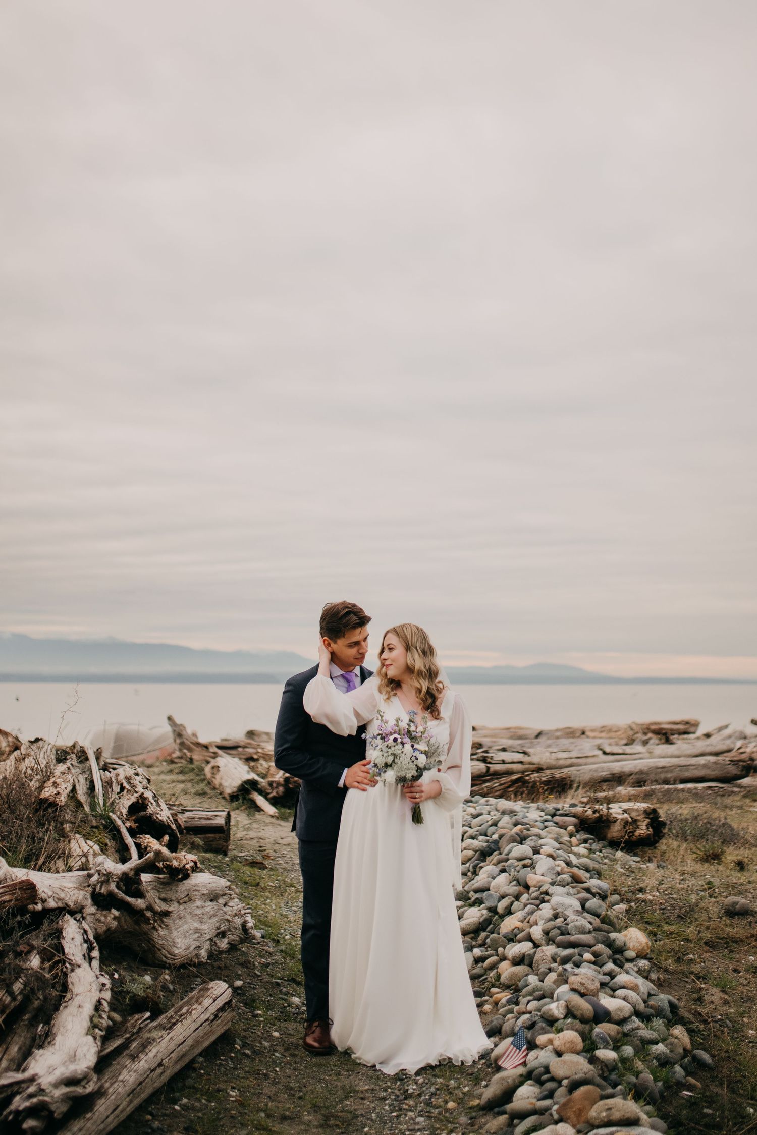 Bride and groom share a romantic moment on a rocky Pacific Northwest beach with driftwood and ocean views.