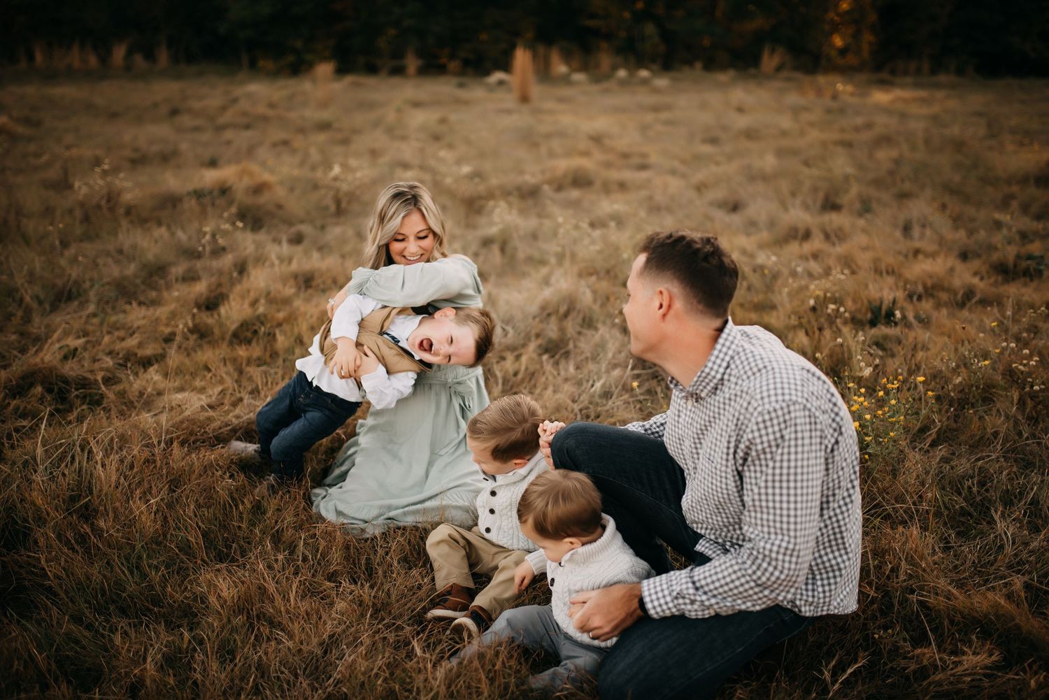 Family of four sitting together in autumn field with parents cuddling young children.