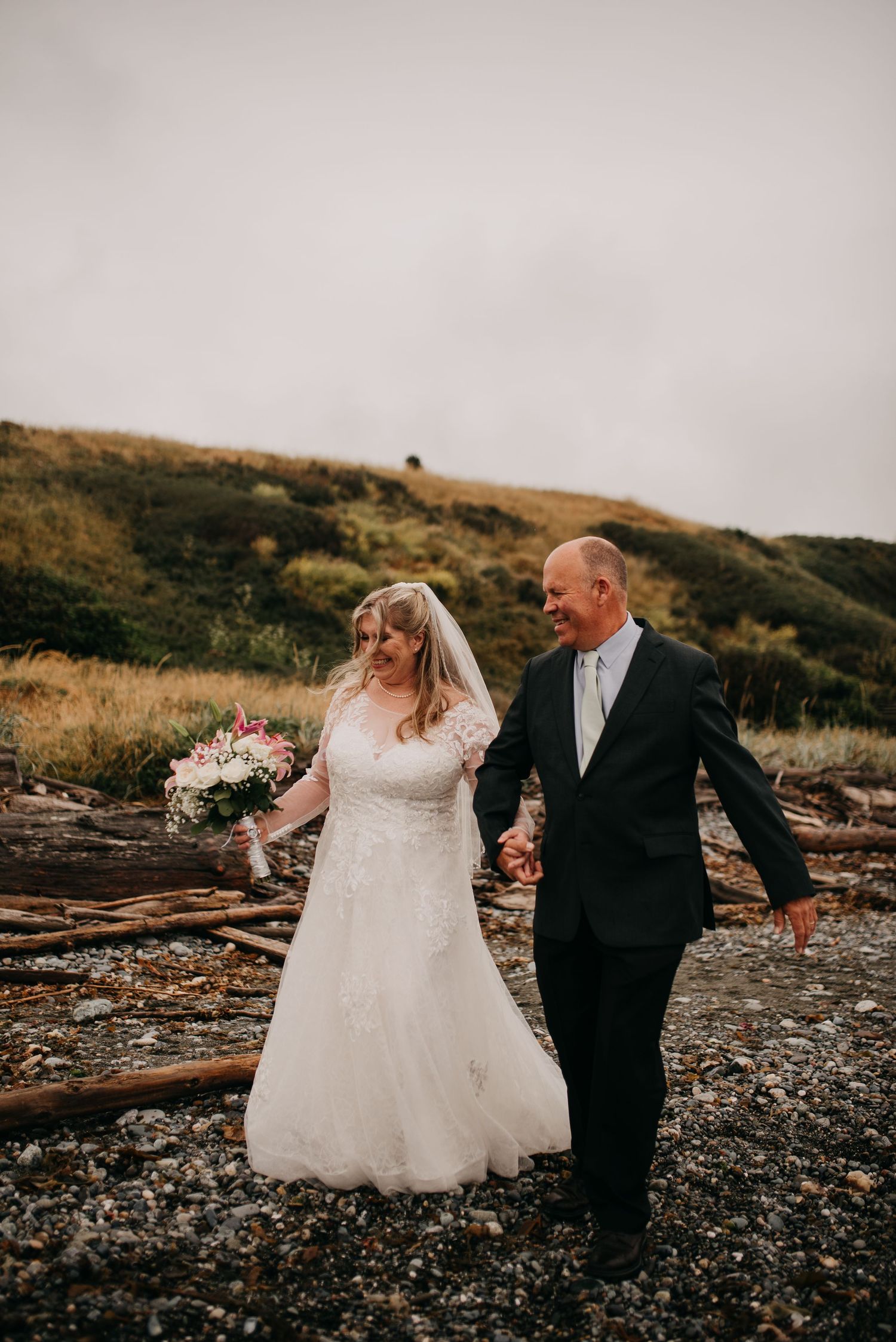 Bride and groom walk hand in hand along a rocky coastal beach with rolling green hills behind them.