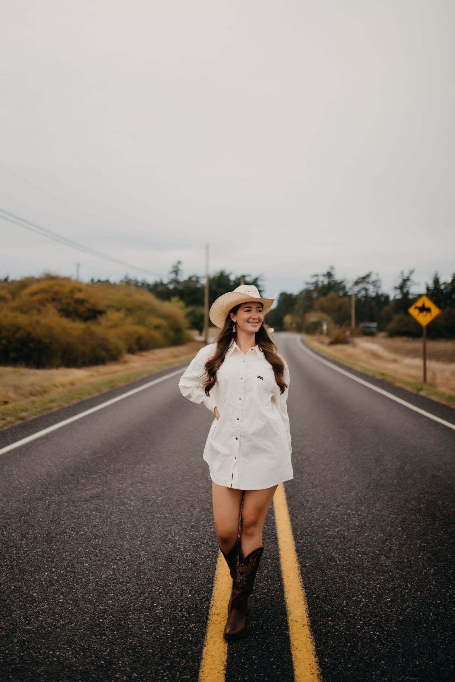 Smiling young woman in white shirt dress and cowboy hat walking on rural road with boots.
