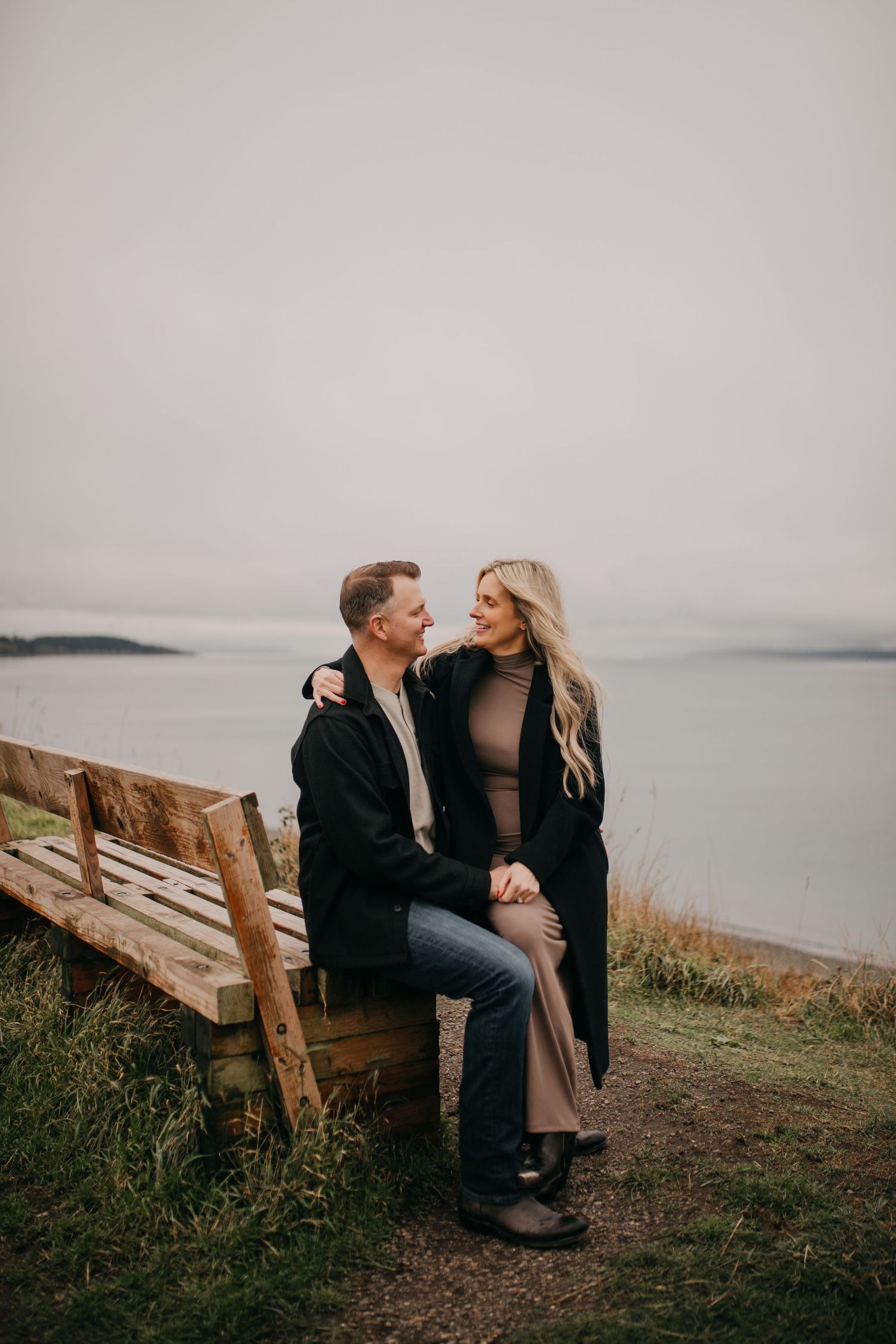 Couple sitting on a rustic wooden bench overlooking a gray ocean, gazing lovingly at each other.