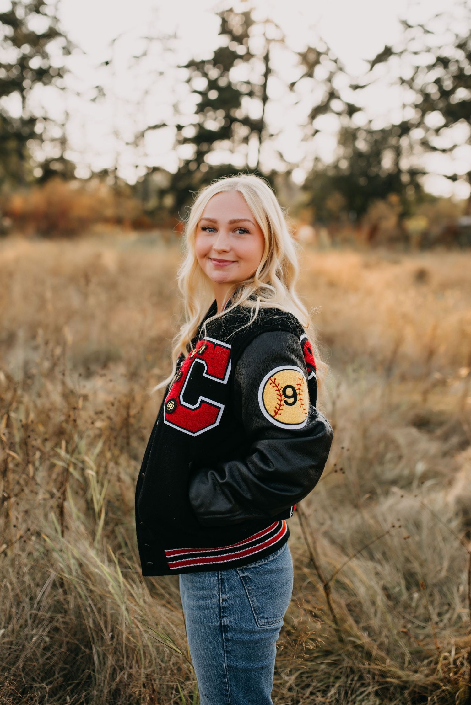 Blonde teen smiles over shoulder wearing varsity letterman jacket in fall field for senior photos.