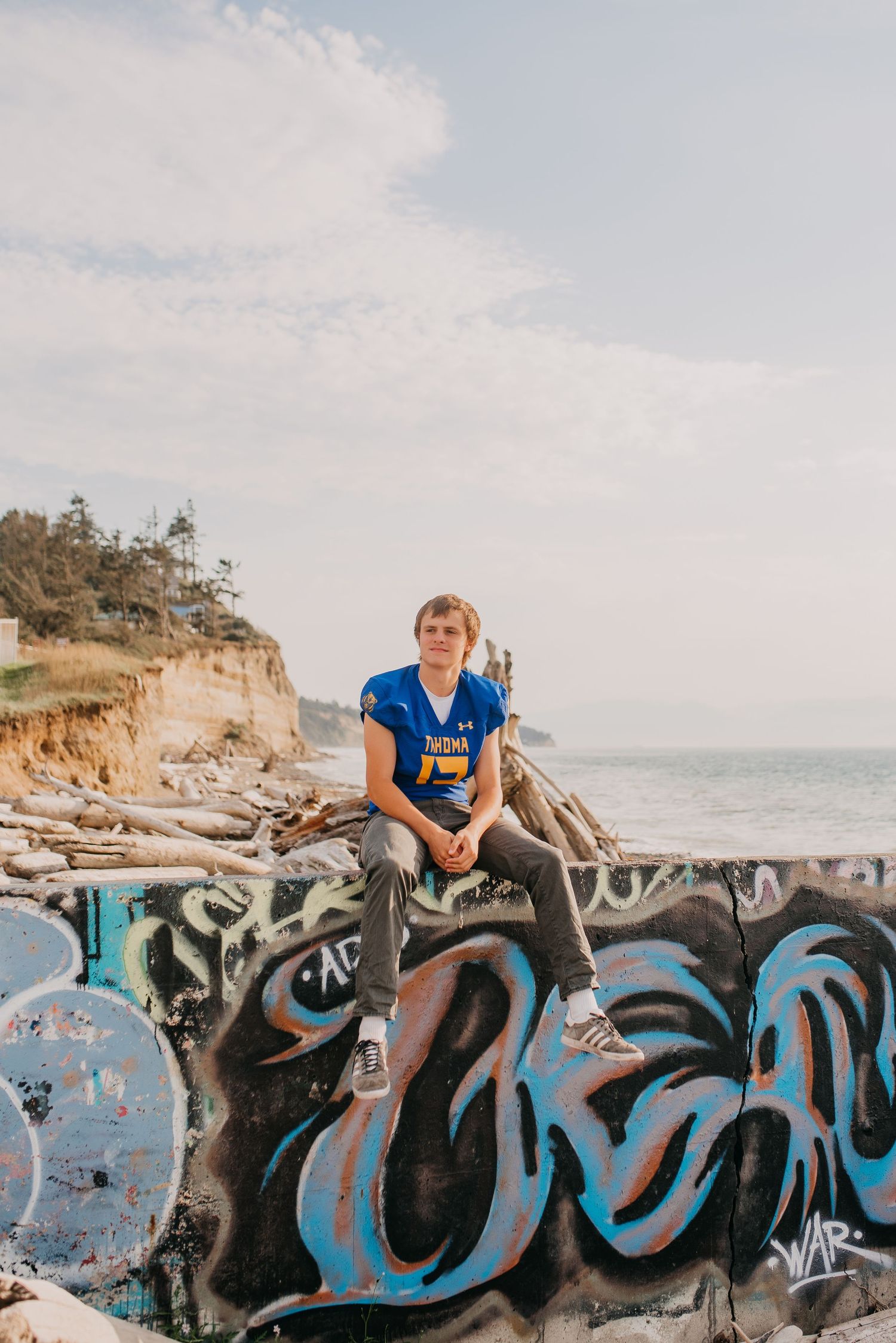 Young man in blue football jersey sits atop graffiti wall with ocean cliffs in background.
