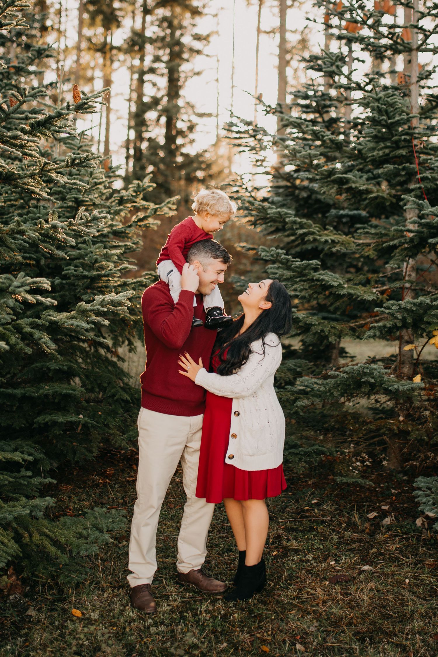 Family of three posing among Christmas trees, father lifting toddler while mother embraces them in festive outfits.