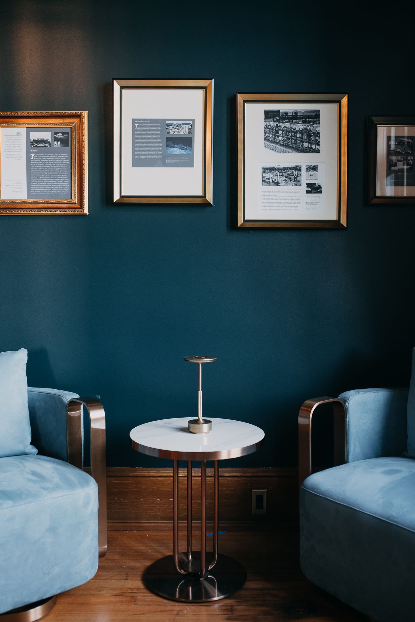 Elegant lounge seating area with two blue velvet chairs, marble side table, and framed artwork on a dark teal wall.