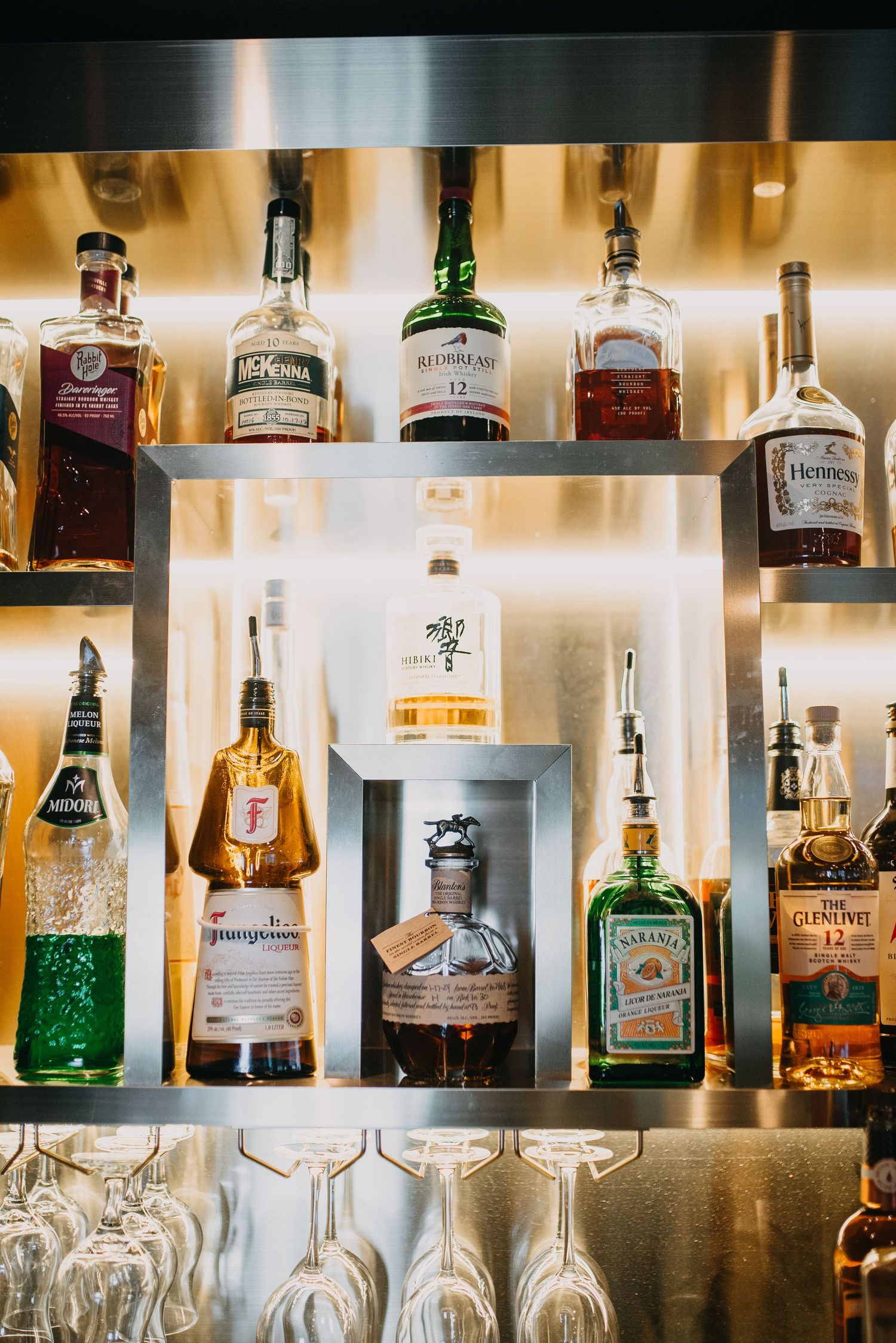 Illuminated bar shelf displaying premium whiskey and spirits bottles with hanging wine glasses below.