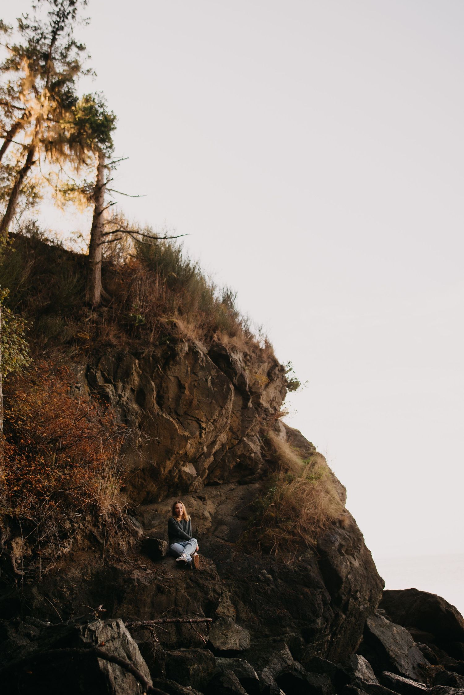 Senior Photography with a girl sitting alone on rocky coastal cliff surrounded by trees and autumn foliage at Rosario Beach in Anacortes.