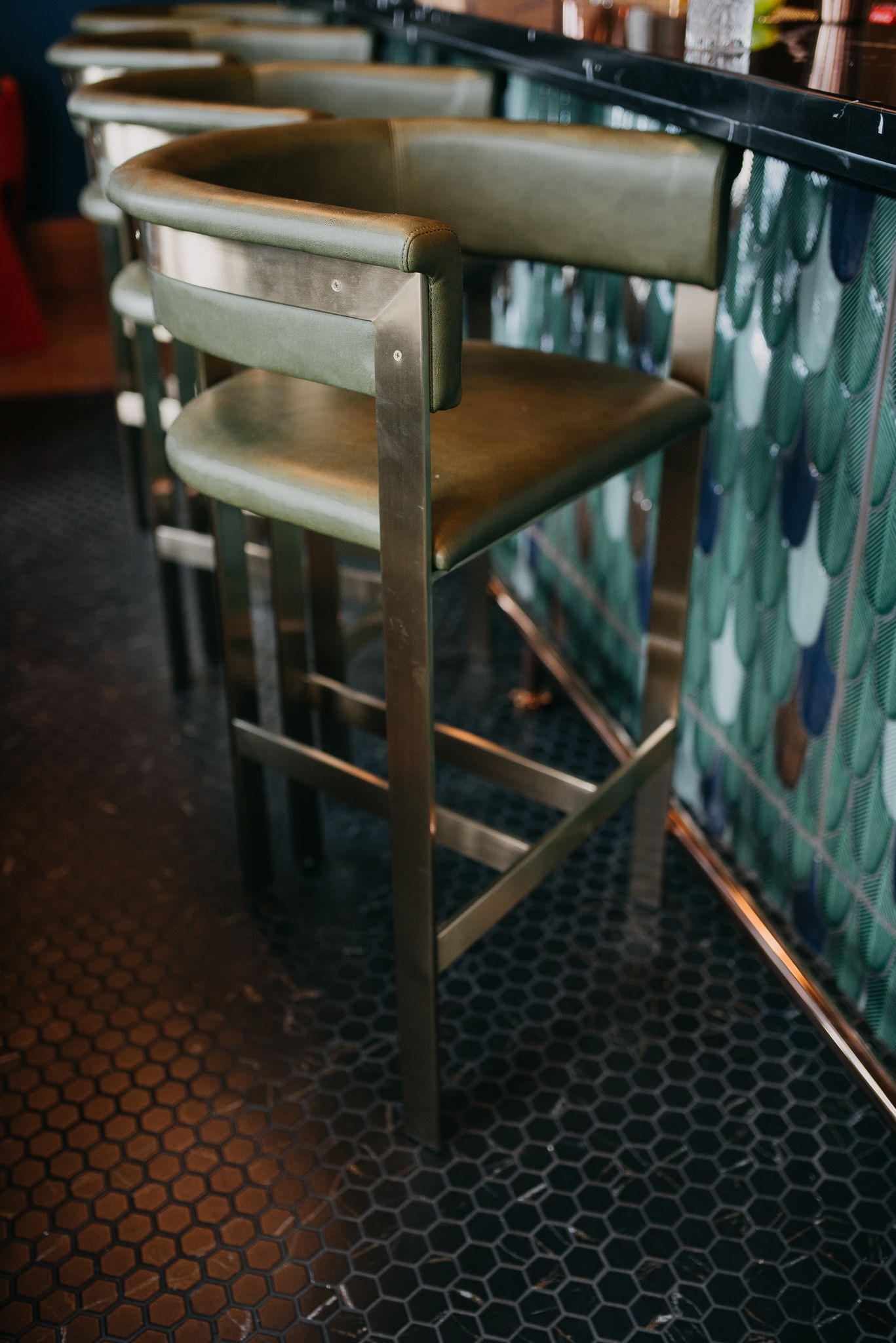 A row of green leather bar stools with metal frames beside a teal mosaic-tiled bar counter.
